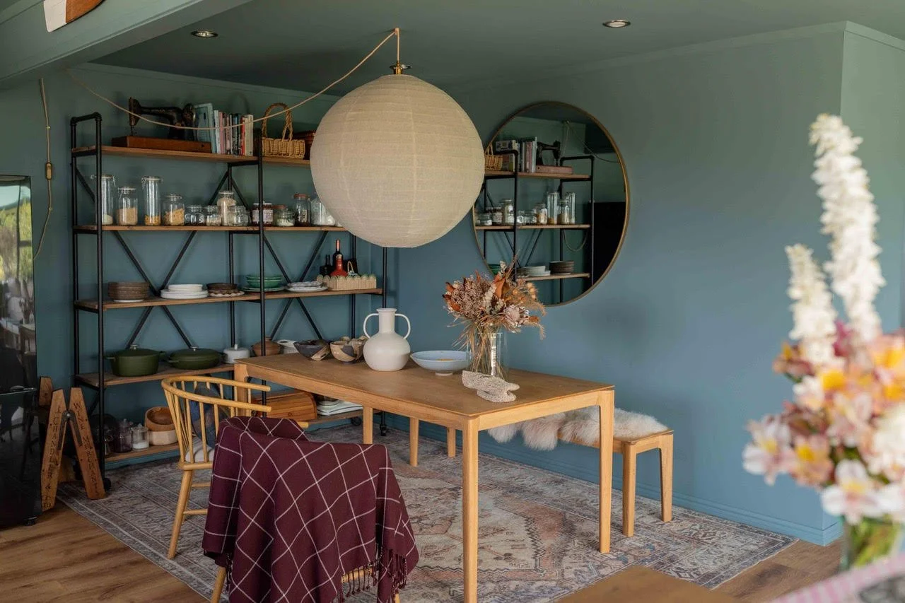 A cozy dining room with a wooden table, patterned rug, and a mix of chairs and benches. Decor includes a white vase, dried flowers, and a large paper lantern hanging from the ceiling. The back wall features open shelves with glass jars, bowls, and bo