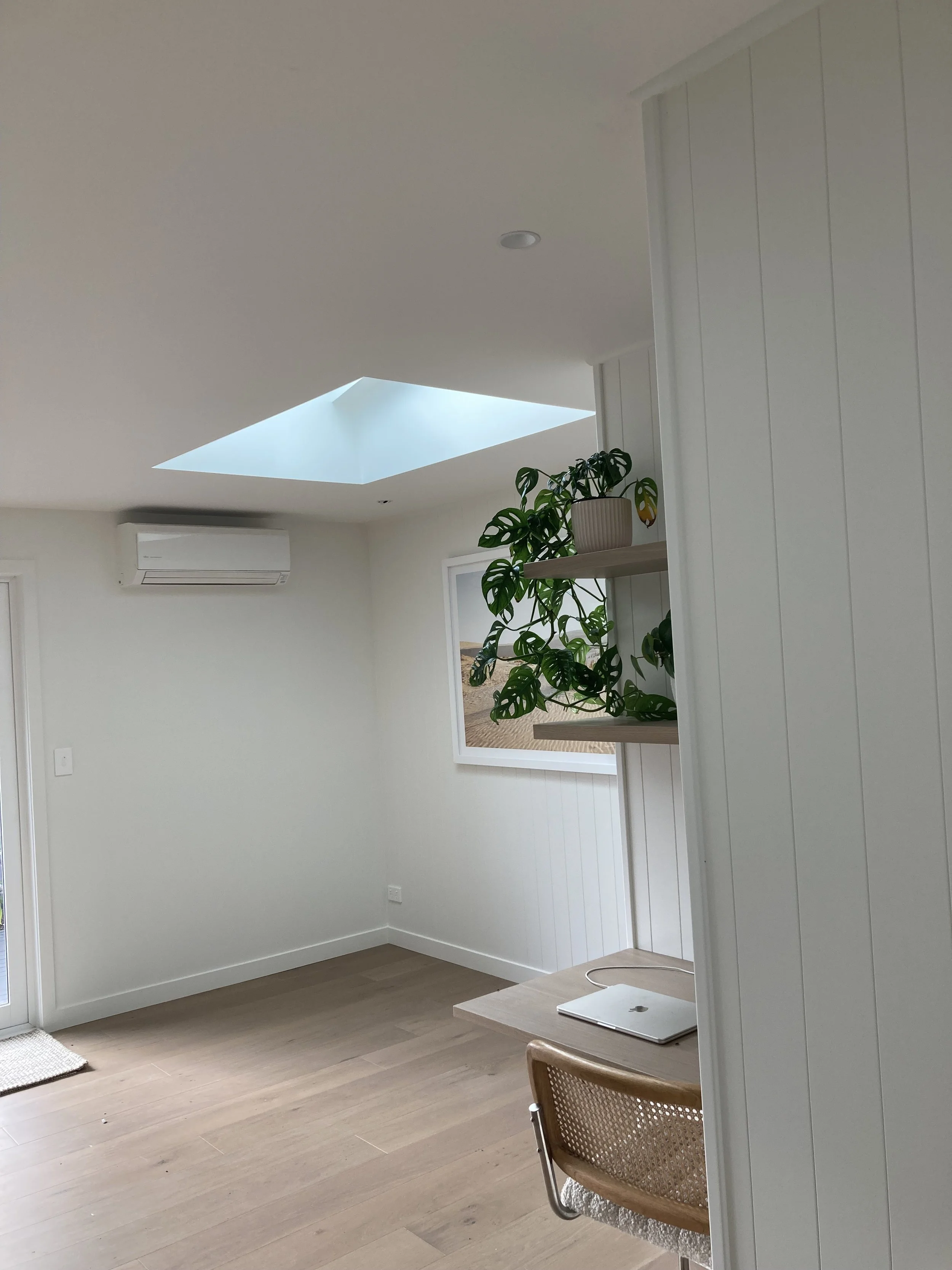 Minimalist room with white walls, skylight, wooden floor, a potted plant on a wall shelf, a window, a laptop on a desk, and a wooden chair.