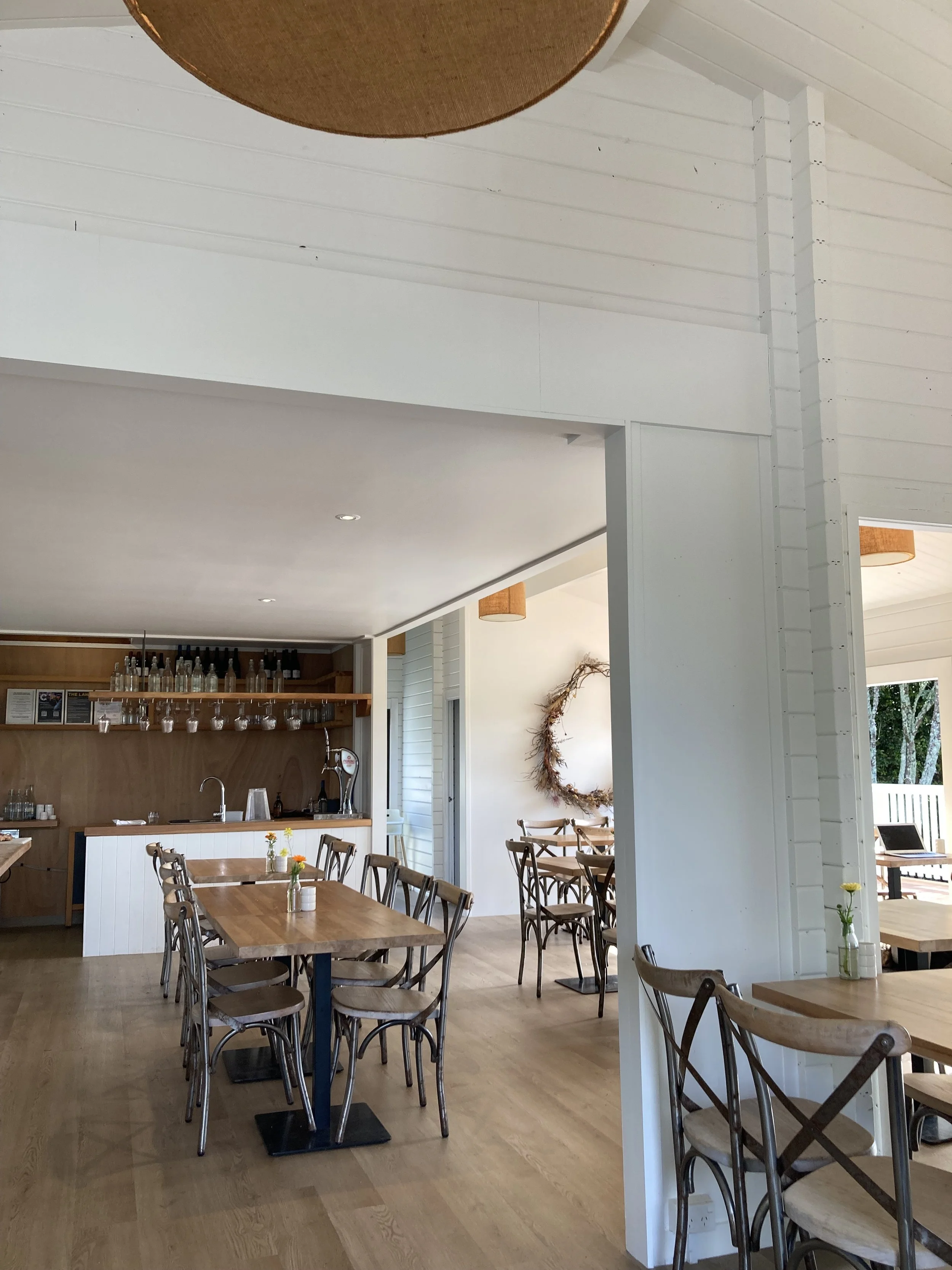 A cozy restaurant or café with wooden tables and chairs, a bar area in the background with glassware and bottles, and natural light coming through windows, decorated with a wreath and small vases of flowers.