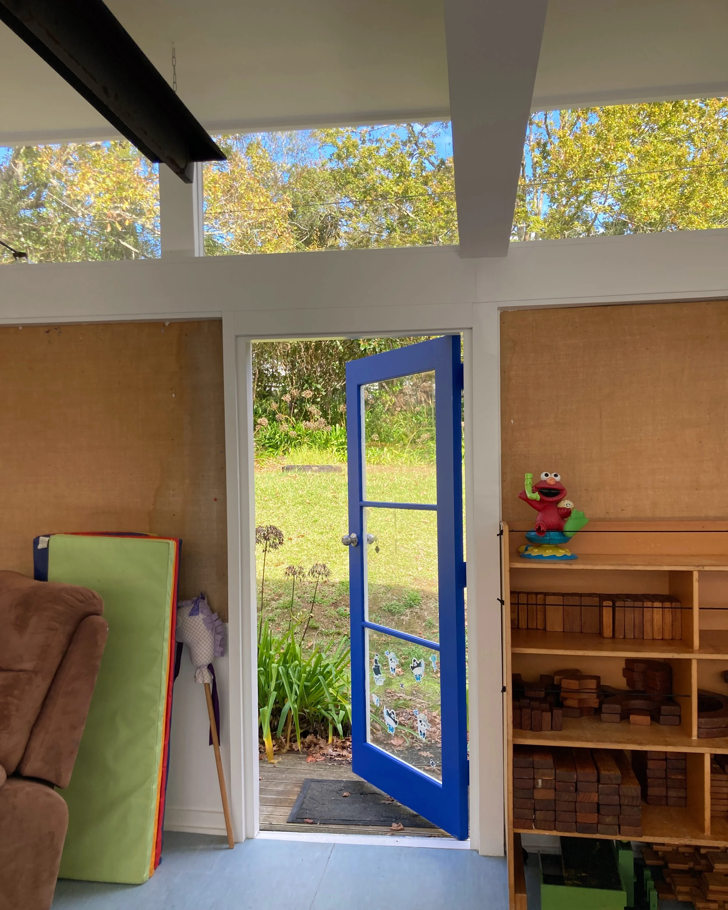 Open blue door leading outdoor garden area with green grass and plants. Indoor room with bookshelf and Sesame Street figure on the right.