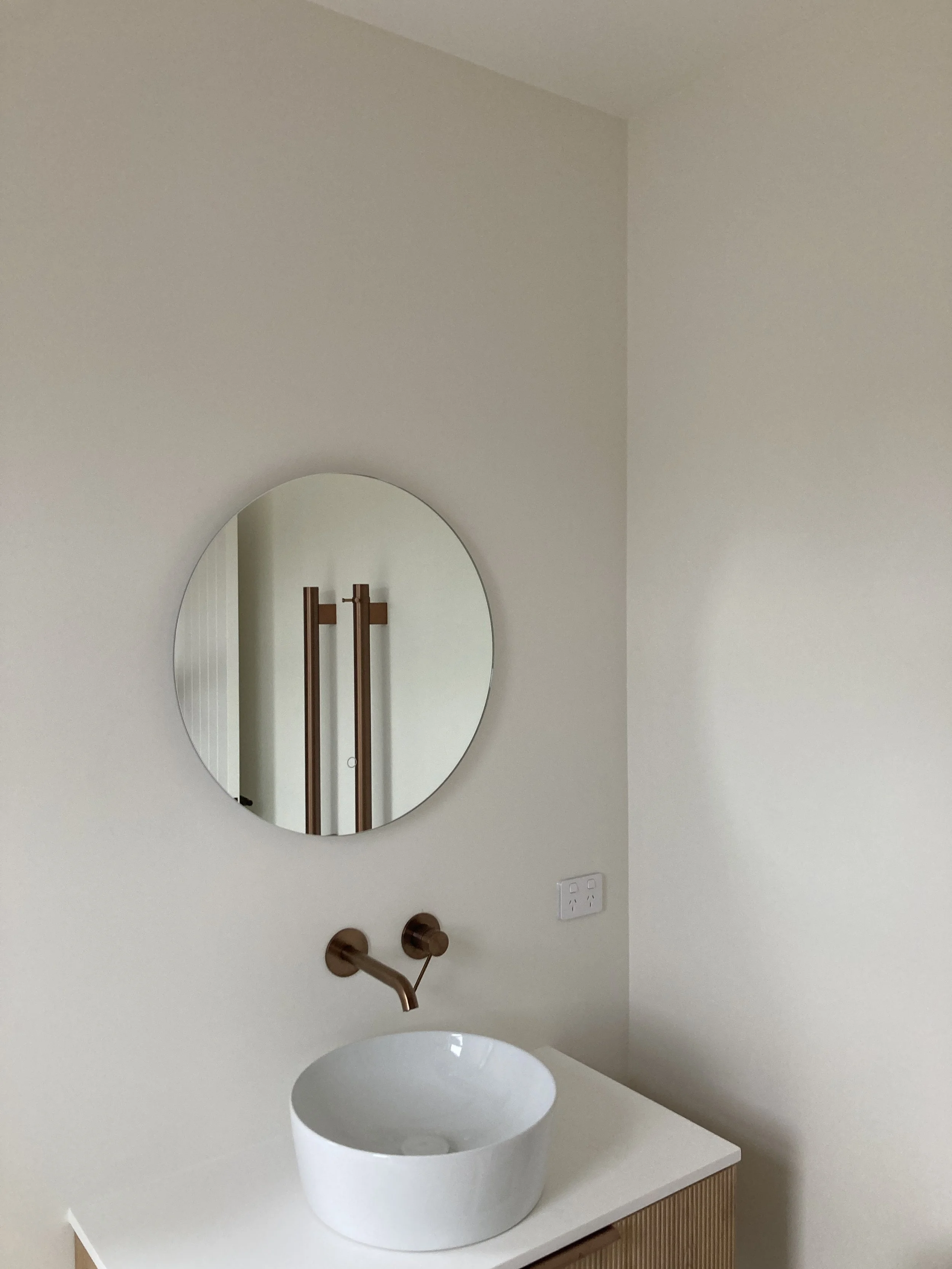 Minimalist bathroom vanity with a white vessel sink, a circular mirror above, wooden cabinet below, and a copper wall-mounted faucet.