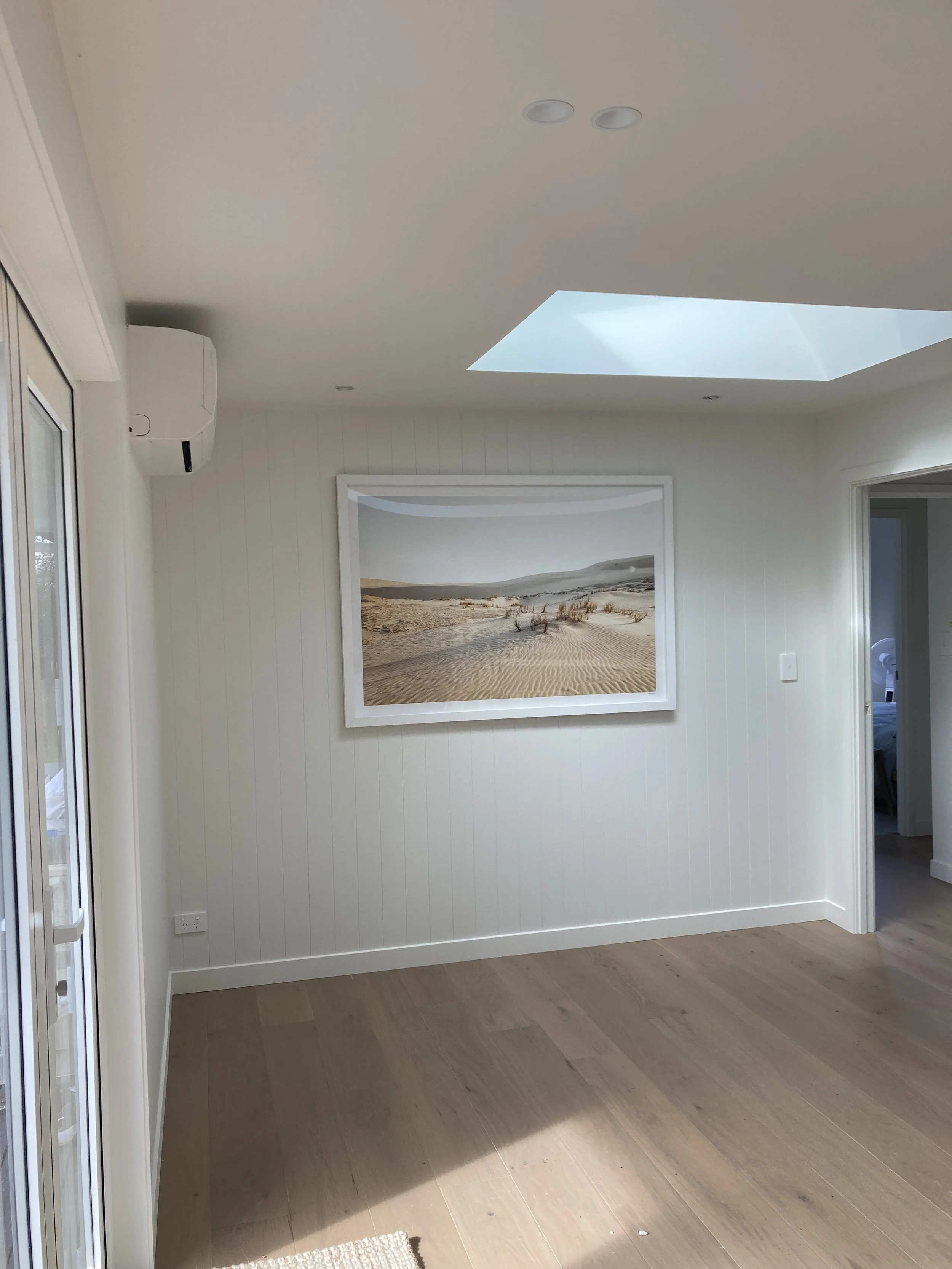 A room with white walls, hardwood flooring, a framed picture of a sandy desert landscape, a skylight, and an air conditioning unit.