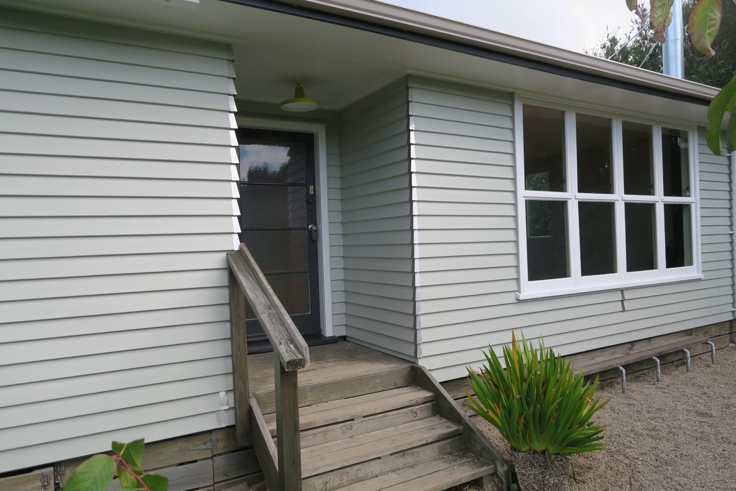Exterior of a house with white siding, a black door with a window, a small wooden ramp, a large window, and a potted plant with long green leaves.