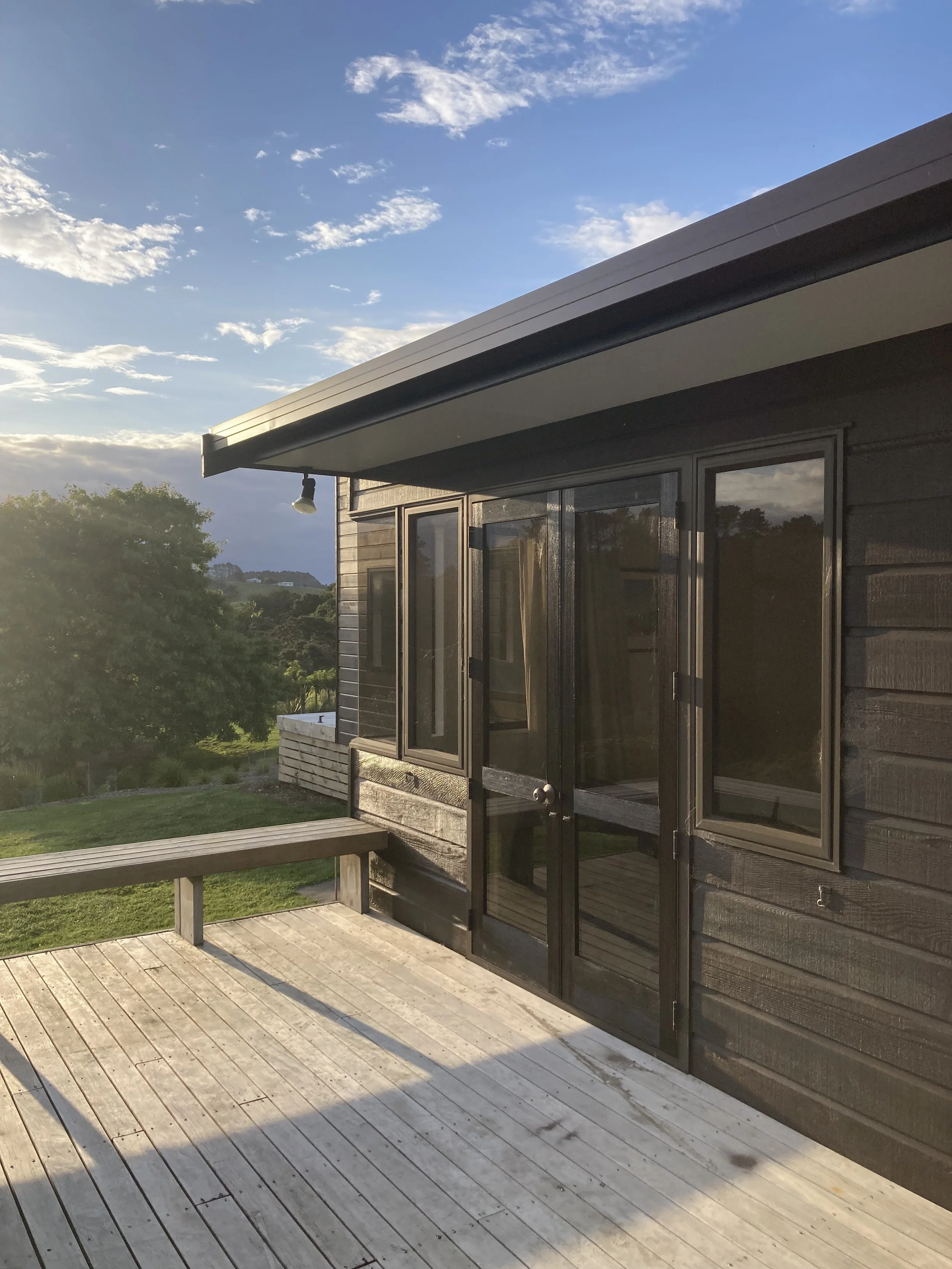 A wooden deck with a bench, adjacent to a black house with large glass doors and windows, overlooking a green landscape with trees and a cloudy sky.