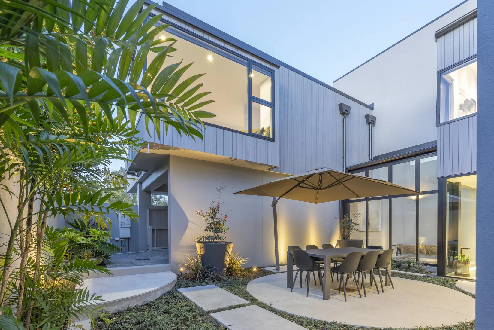 Modern backyard patio with a dining table, chairs, a large umbrella, and potted plants, surrounded by a contemporary house with large windows and outdoor lighting at dusk.