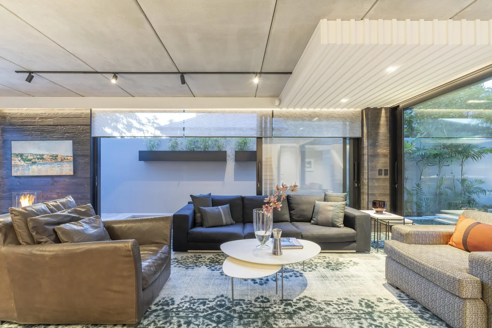 Modern living room with leather and fabric sofas, a patterned rug, and a white coffee table with a large vase of pink flowers, in front of large glass windows with greenery outside.
