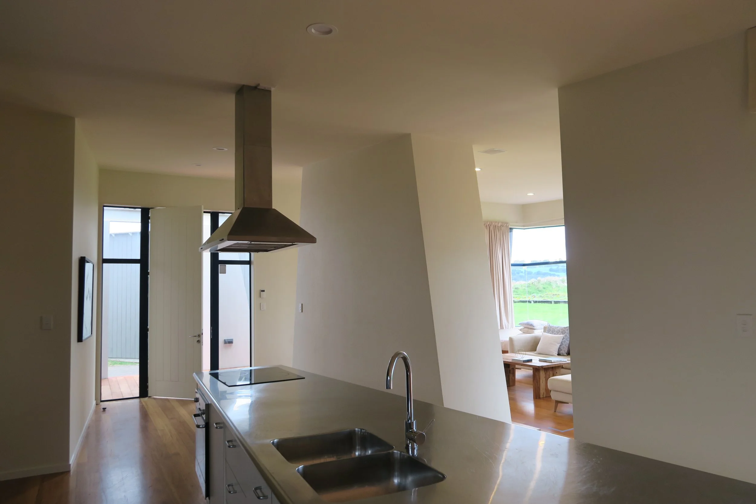 Interior view of a modern kitchen with a sink, stovetop, and a hanging range hood, opening into a living room with large windows and outdoor scenery.