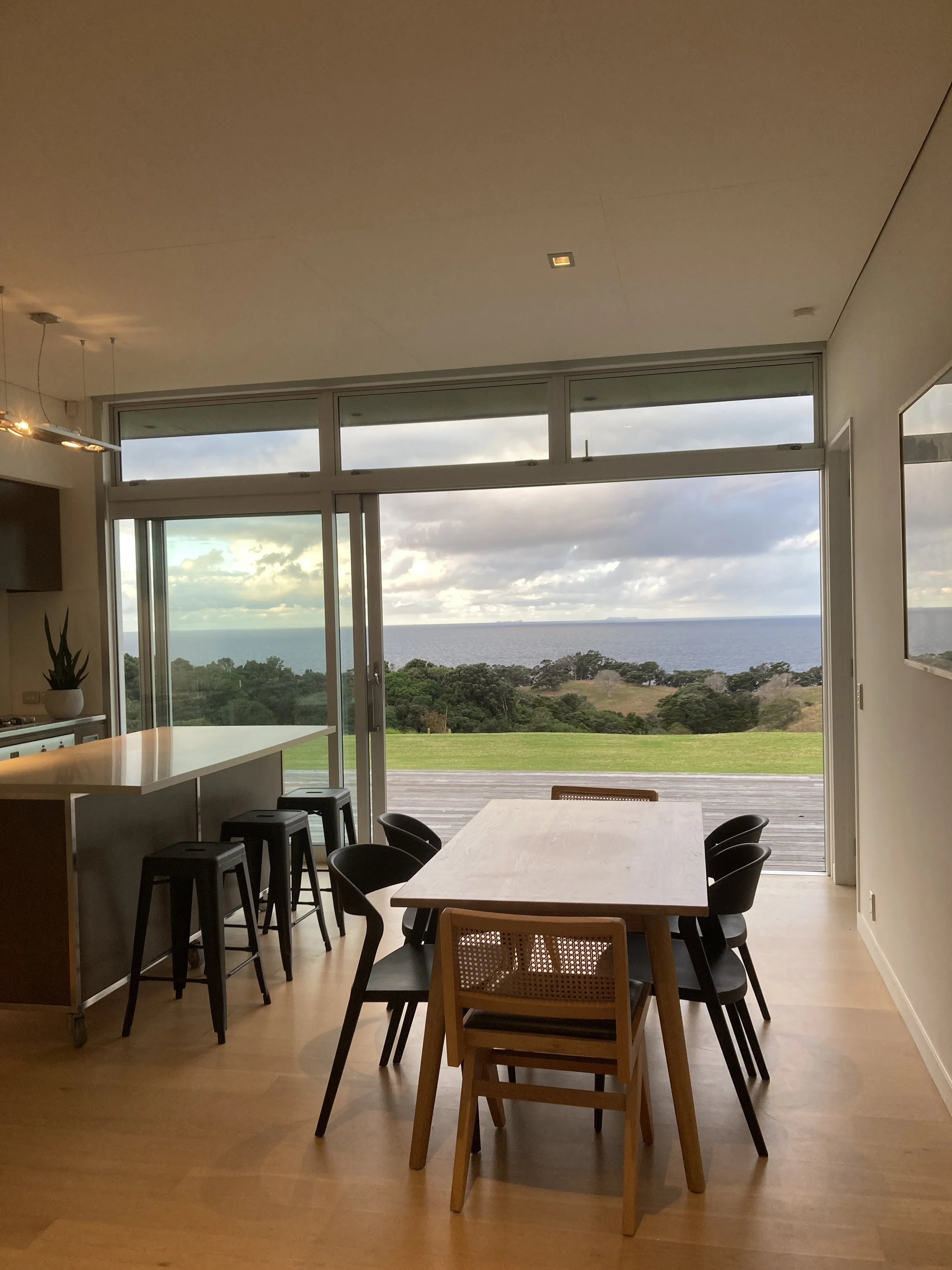 Modern dining area with a wooden table and chairs in front of large glass doors leading outdoors to a green landscape and water view.