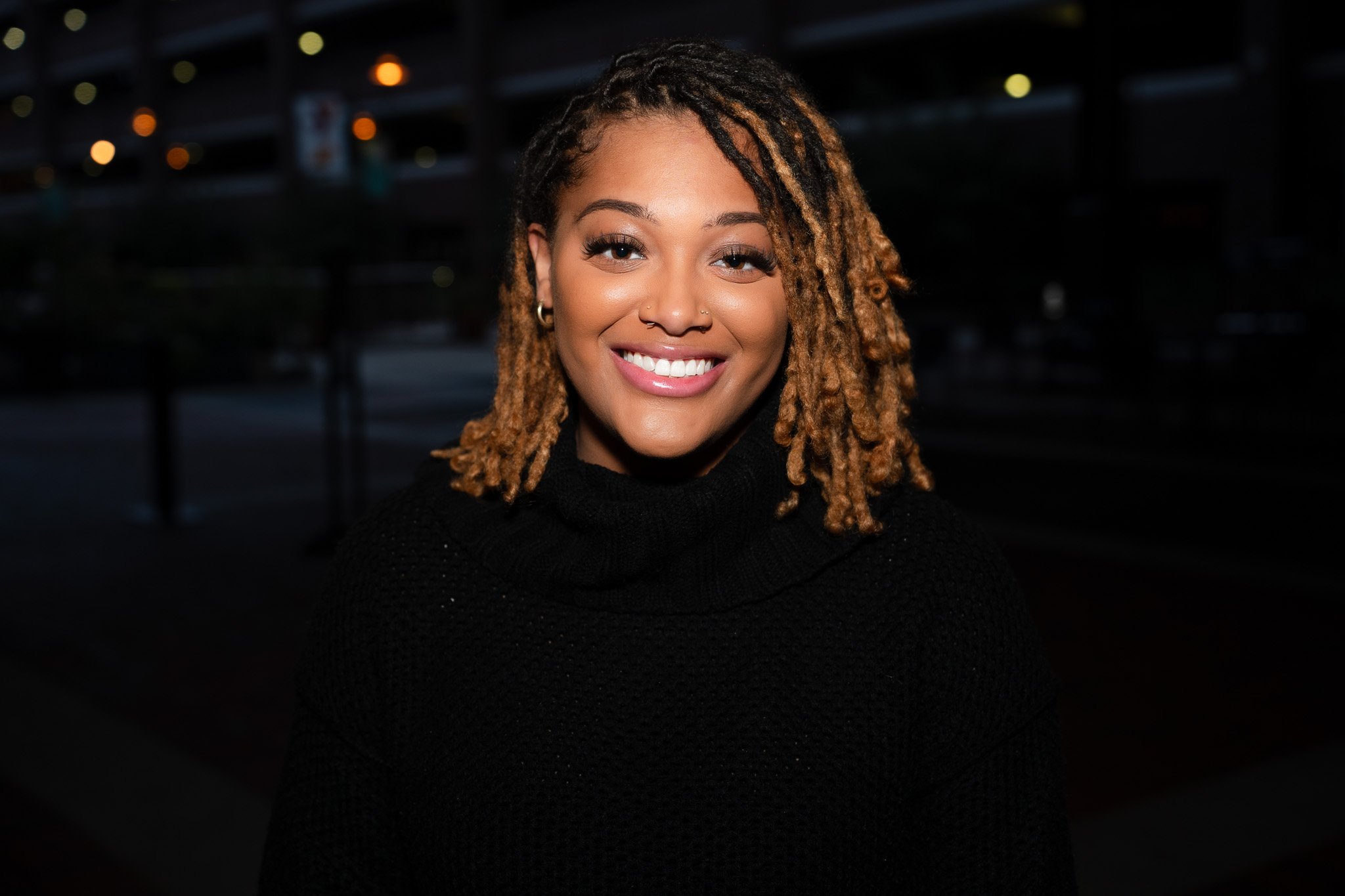 Smiling person with locs wearing a black sweater, standing outdoors at night.