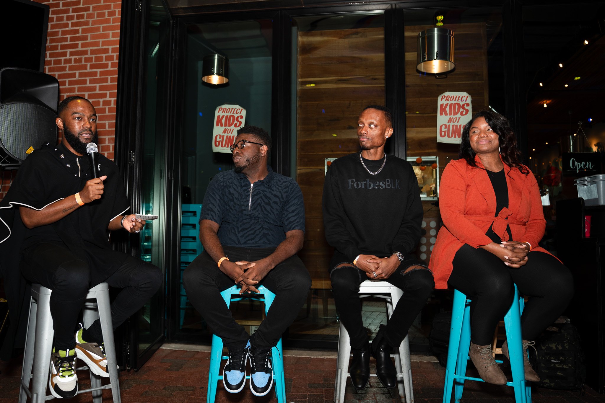 Panel discussion with four people seated on stools outside a venue, with a "Protect Kids Not Guns" sign in the background.