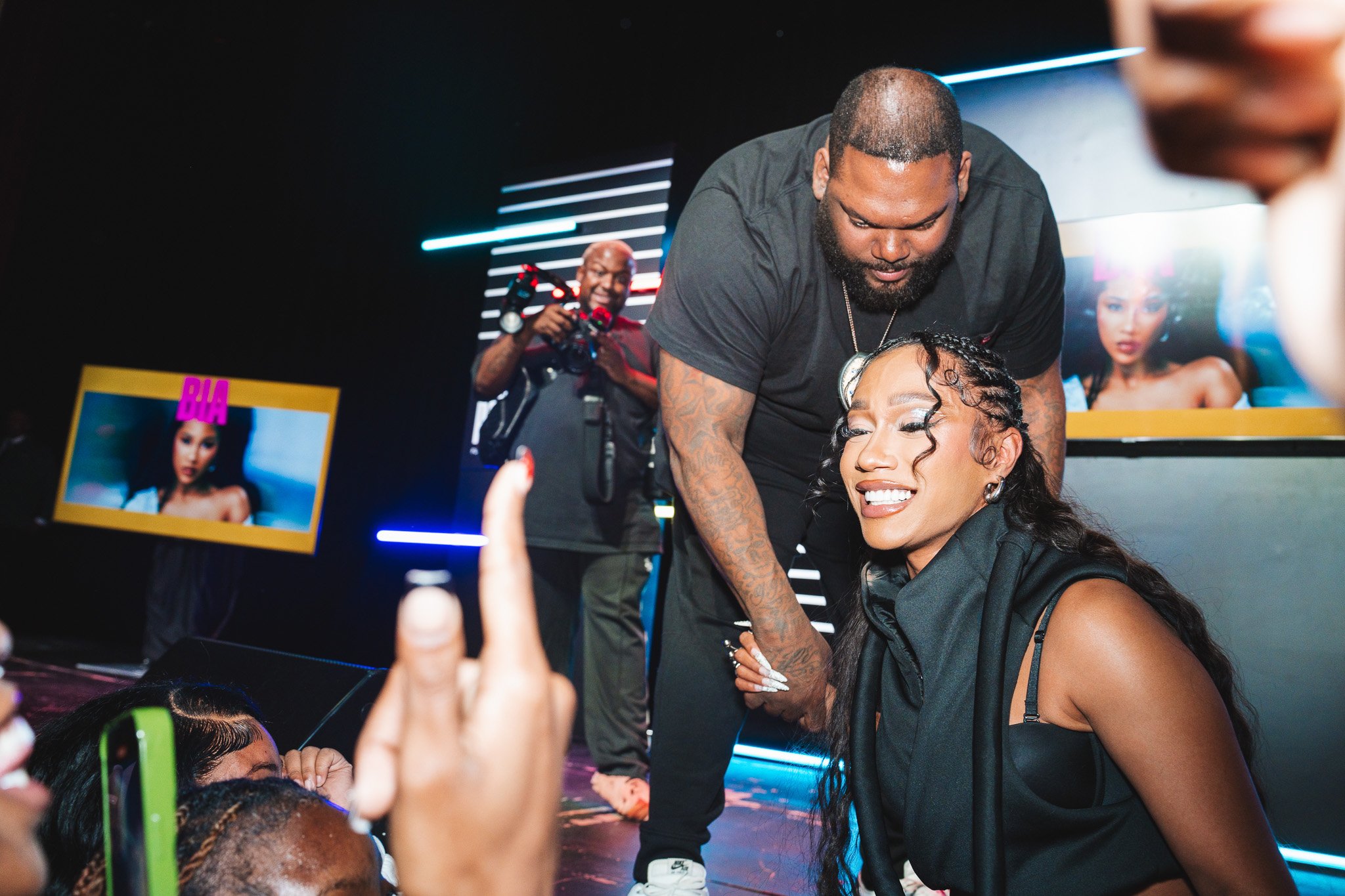 A performer interacting with fans on stage, holding hands and smiling. A person in black operates a camera in the background. A screen displays an image with the text "BIA."