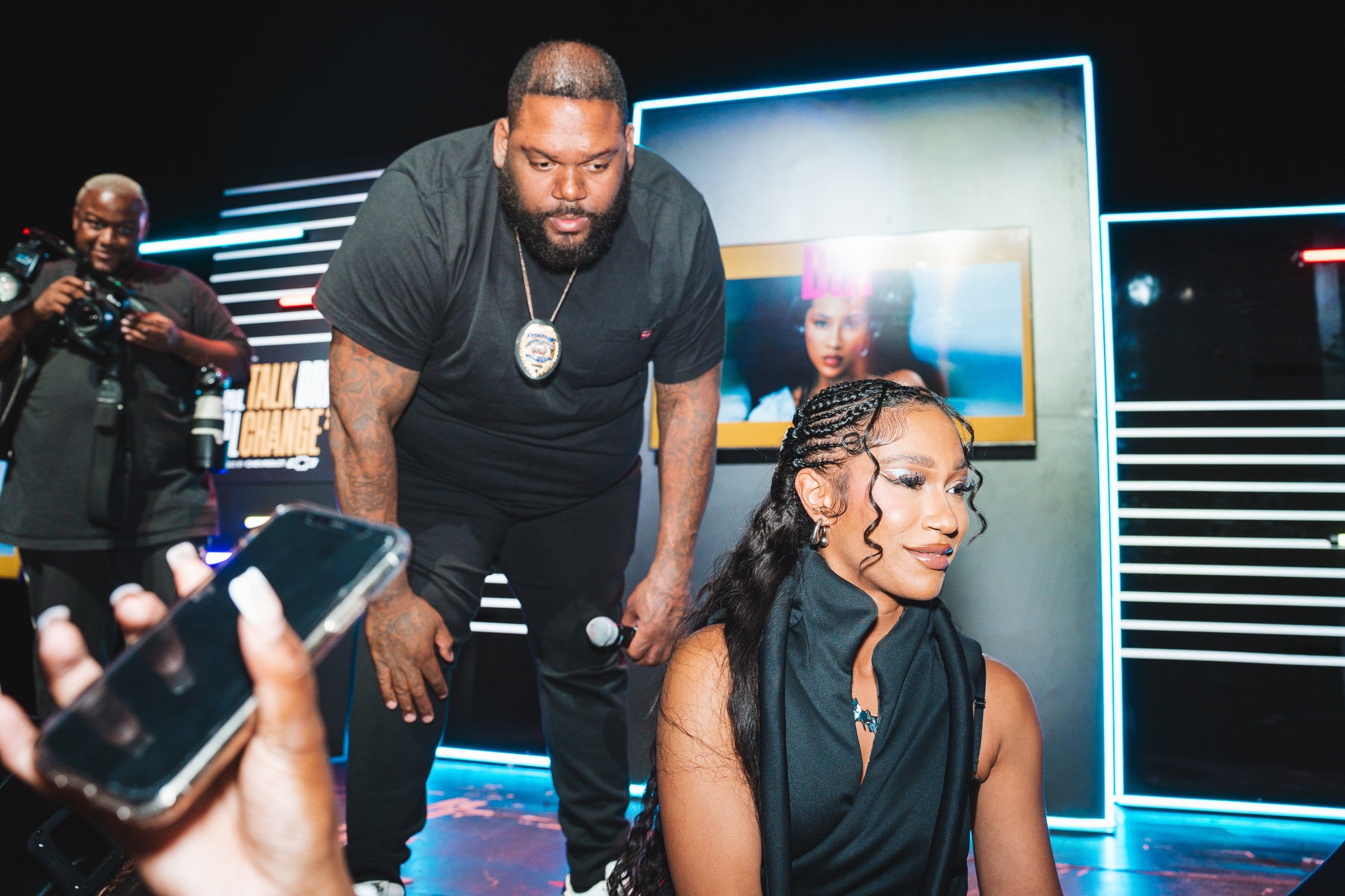 Man and woman in an event setting with neon lights, man holding a microphone, woman sitting with braided hairstyle, a photographer in the background, and a phone in the foreground.