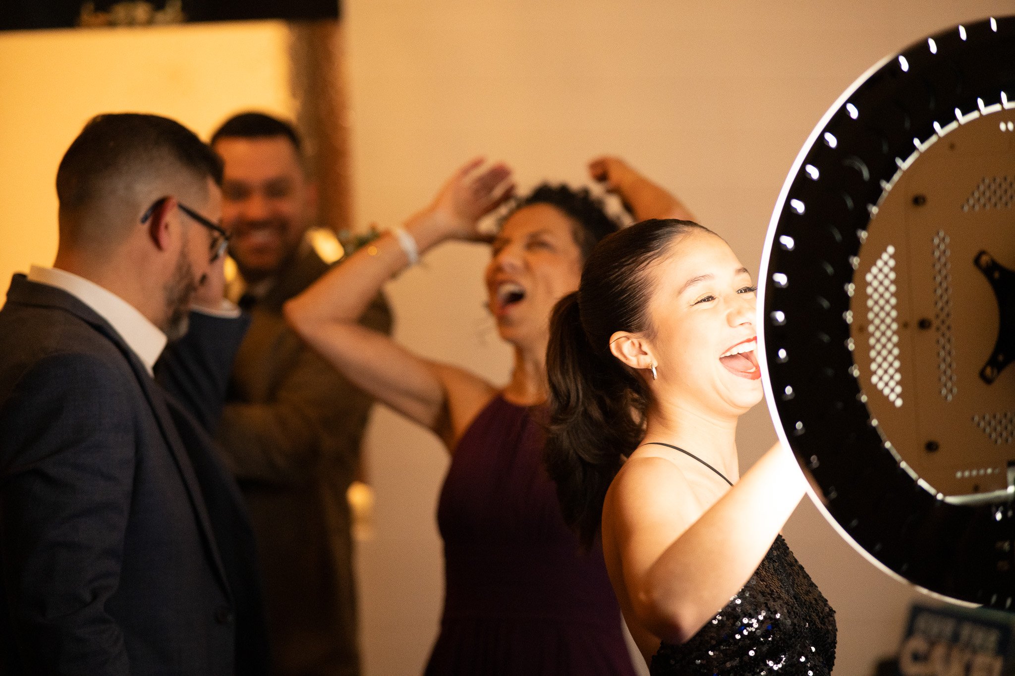 People laughing and posing for a photo booth at an event, with a woman in a black sequin dress in the foreground.