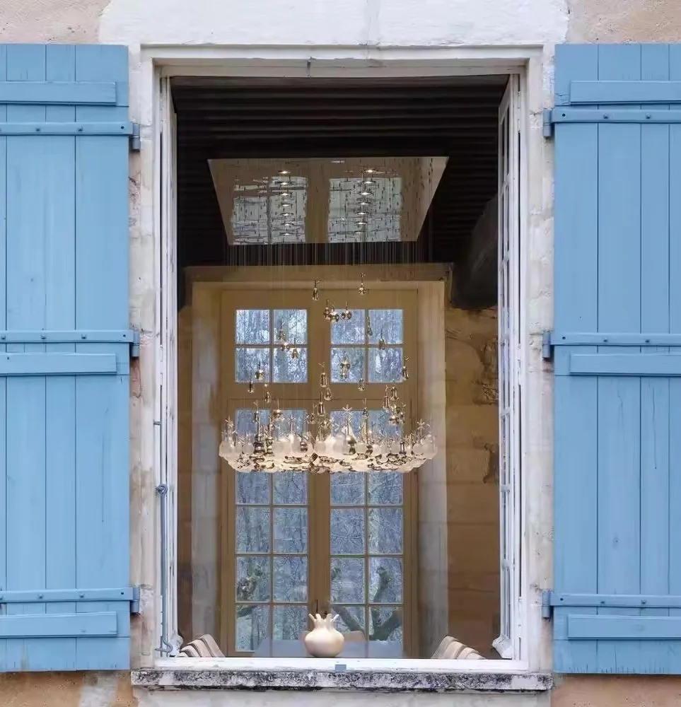 View through an open window with blue shutters showing an elegant chandelier and large windows inside a room.