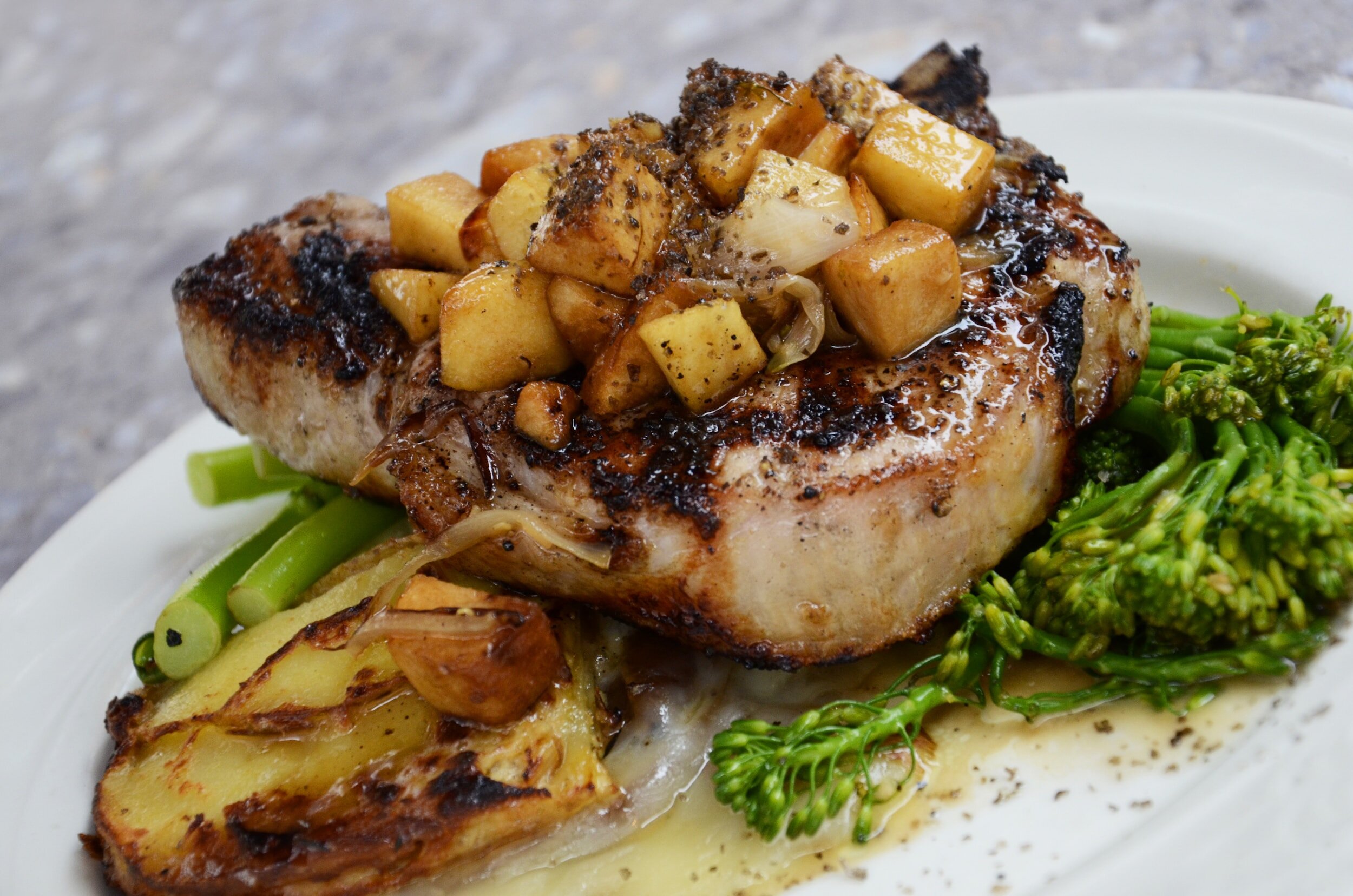 Côte de porc grillée garnie de dés de pommes et de pommes de terre rôties, servie avec des légumes verts comme le brocoli, sur une assiette blanche.
