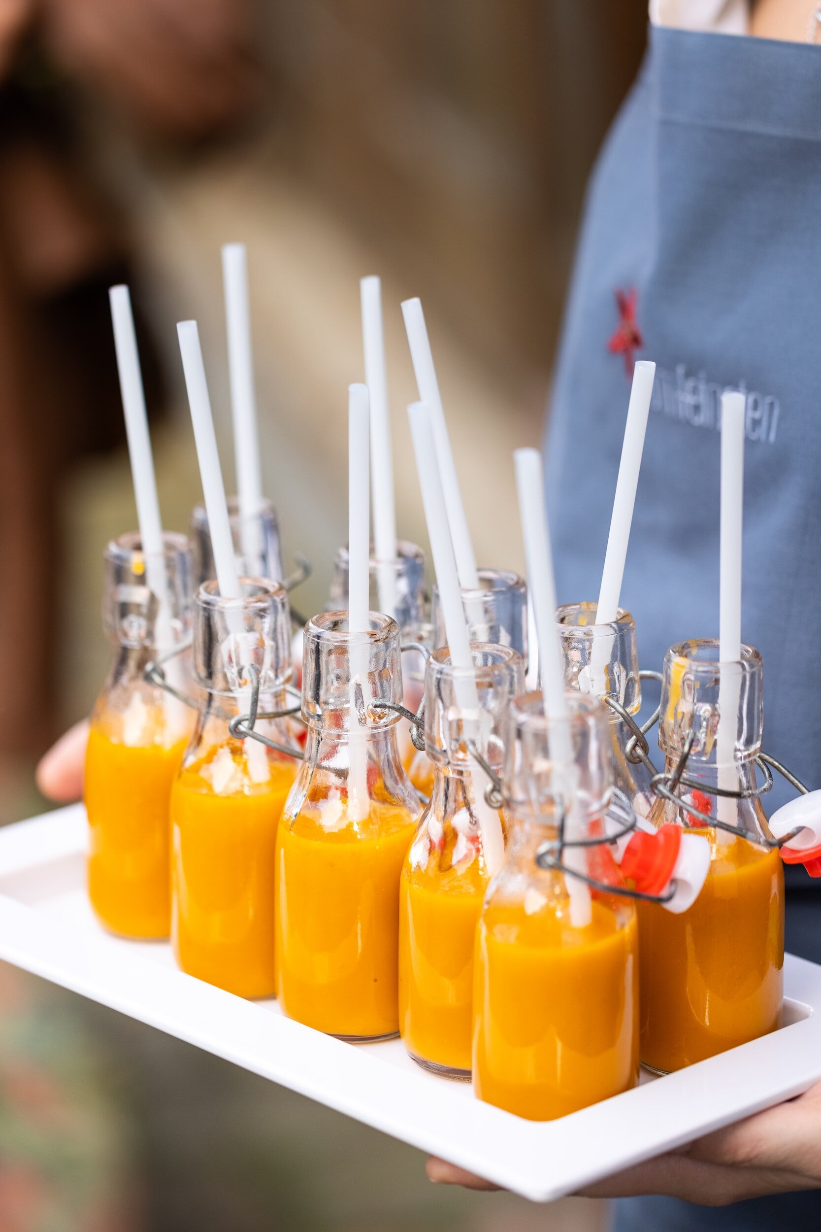 Plateau de petites bouteilles en verre contenant une boisson orange et dotées de pailles blanches, tenu par une personne en tablier.