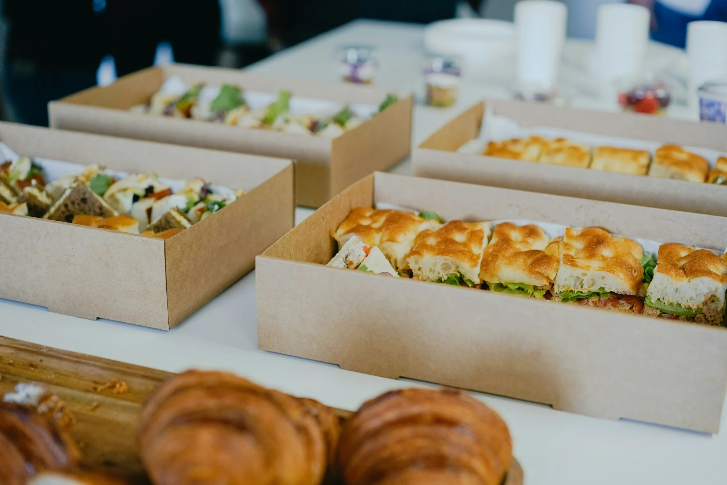 Assortiment de sandwiches, quiches ou cakes salés dans des boîtes en carton sur une table, avec d'autres petits desserts en arrière-plan.