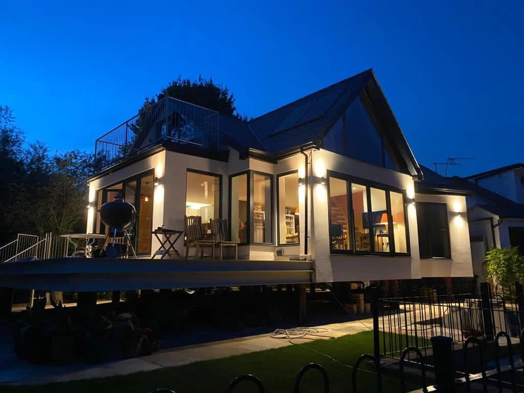 Modern house with large glass windows illuminated at dusk, featuring a deck with outdoor furniture and a grill.