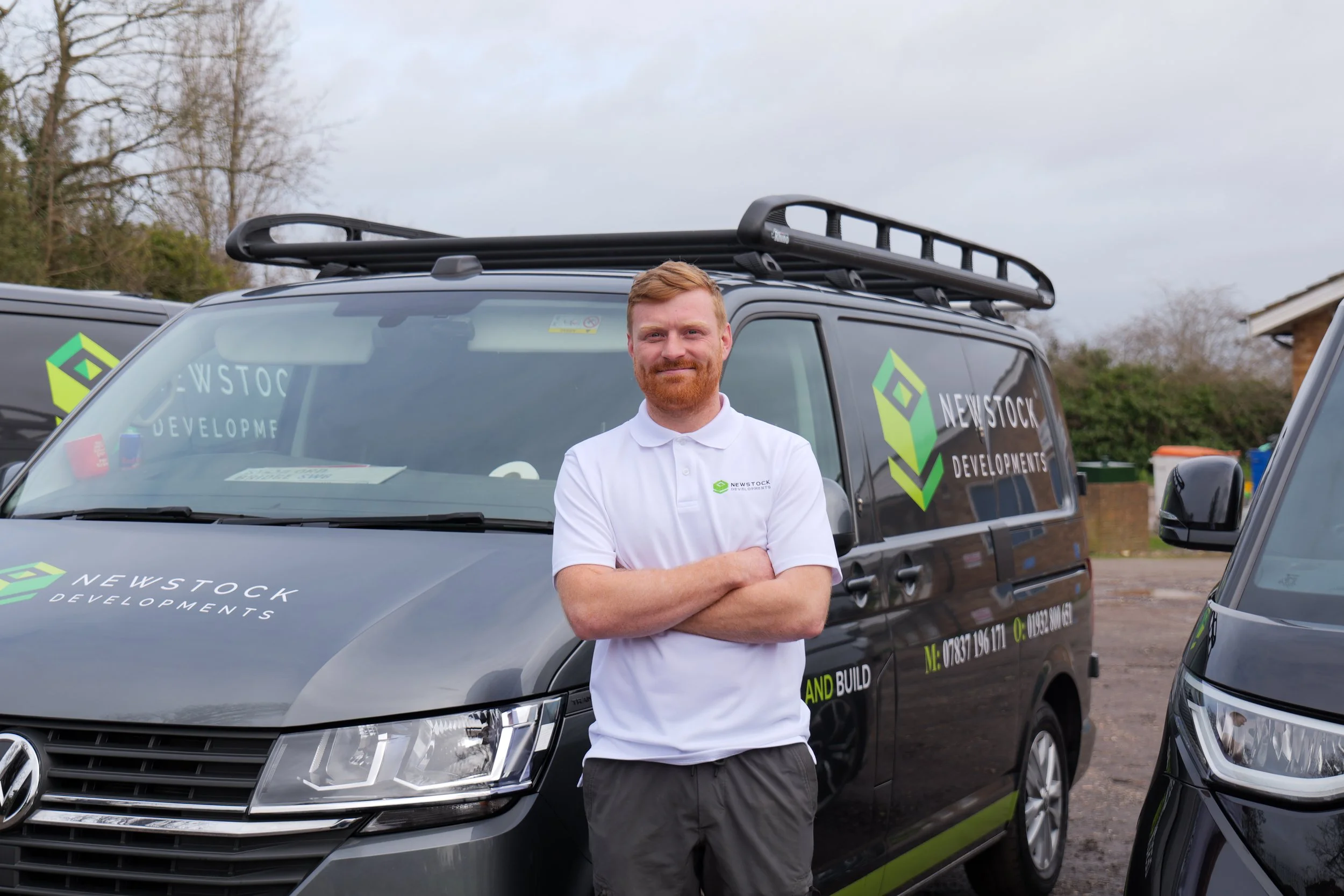 A man with red hair and beard standing in front of a black Newstock Developments van with his arms crossed, wearing a white polo shirt, outdoors on a cloudy day.