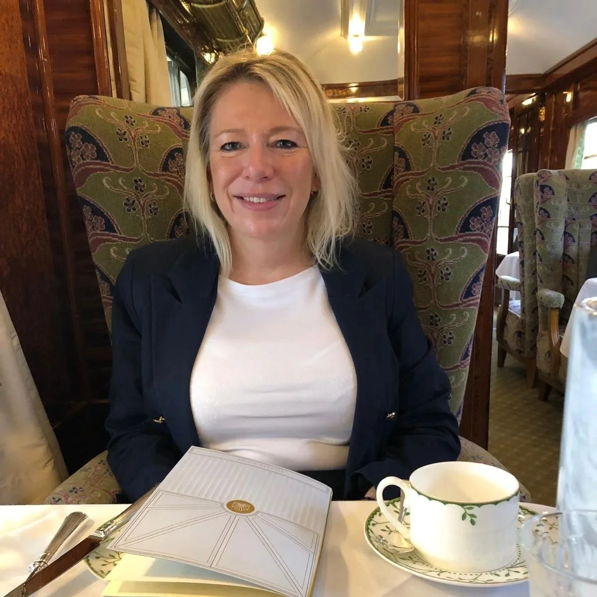 A woman with blonde hair smiling, sitting at a dining table in a restaurant, wearing a white top and a dark blazer, with a menu and a teacup in front of her.