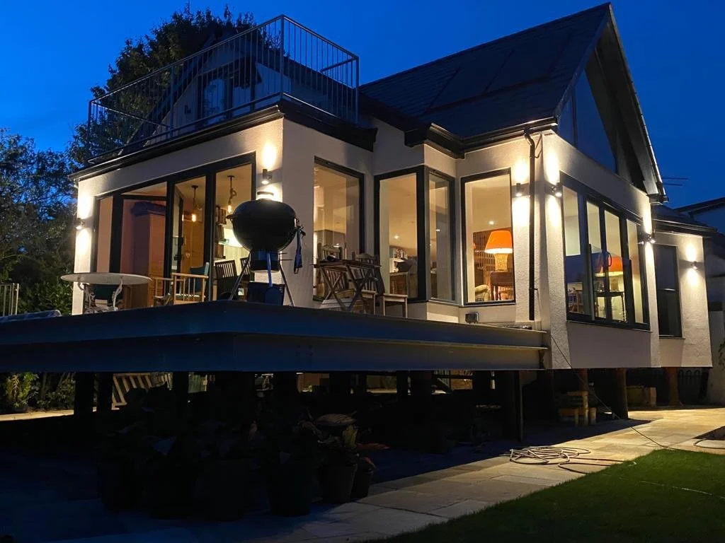 A two-story house illuminated at night with large glass windows and a balcony on the roof, outdoor furniture, and a grill on the patio.
