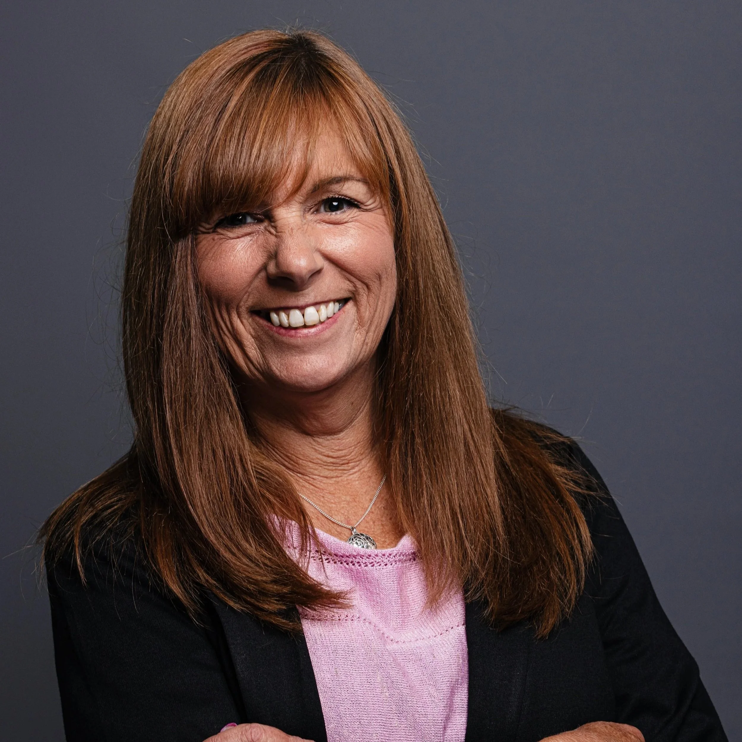 A woman with long dark hair, smiling, wearing a black blazer over a pink sweater and a necklace, against a dark grey background.