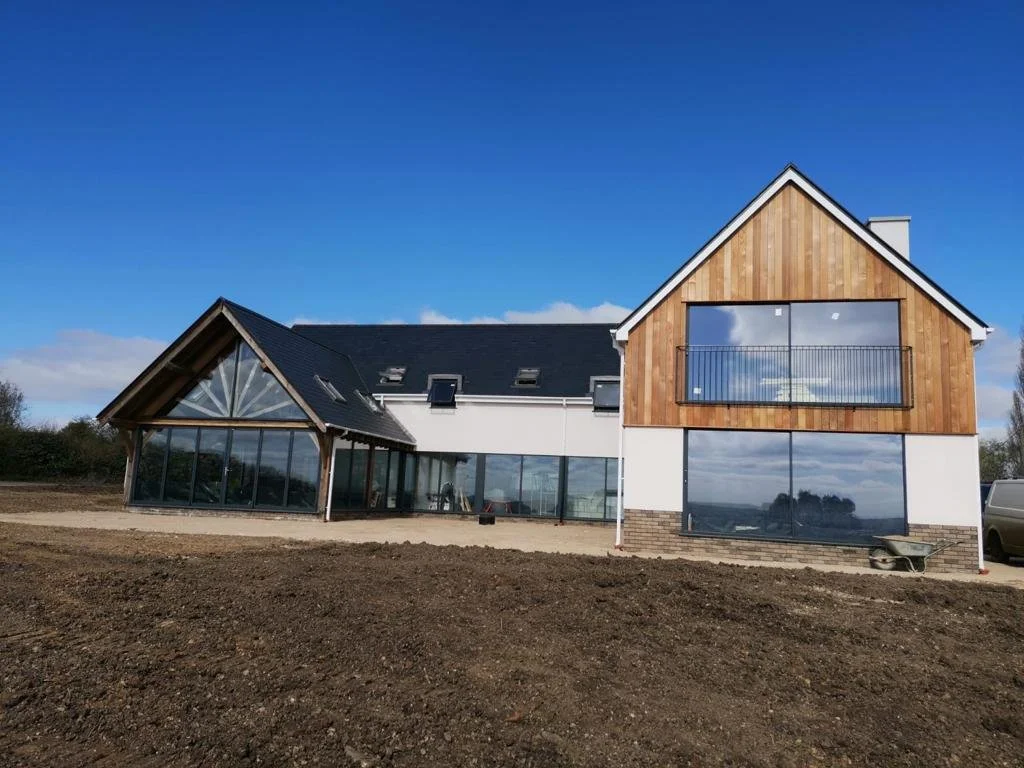 A modern house with large glass windows, a sloped black roof, and wooden paneling on part of the exterior, set against a bright blue sky.