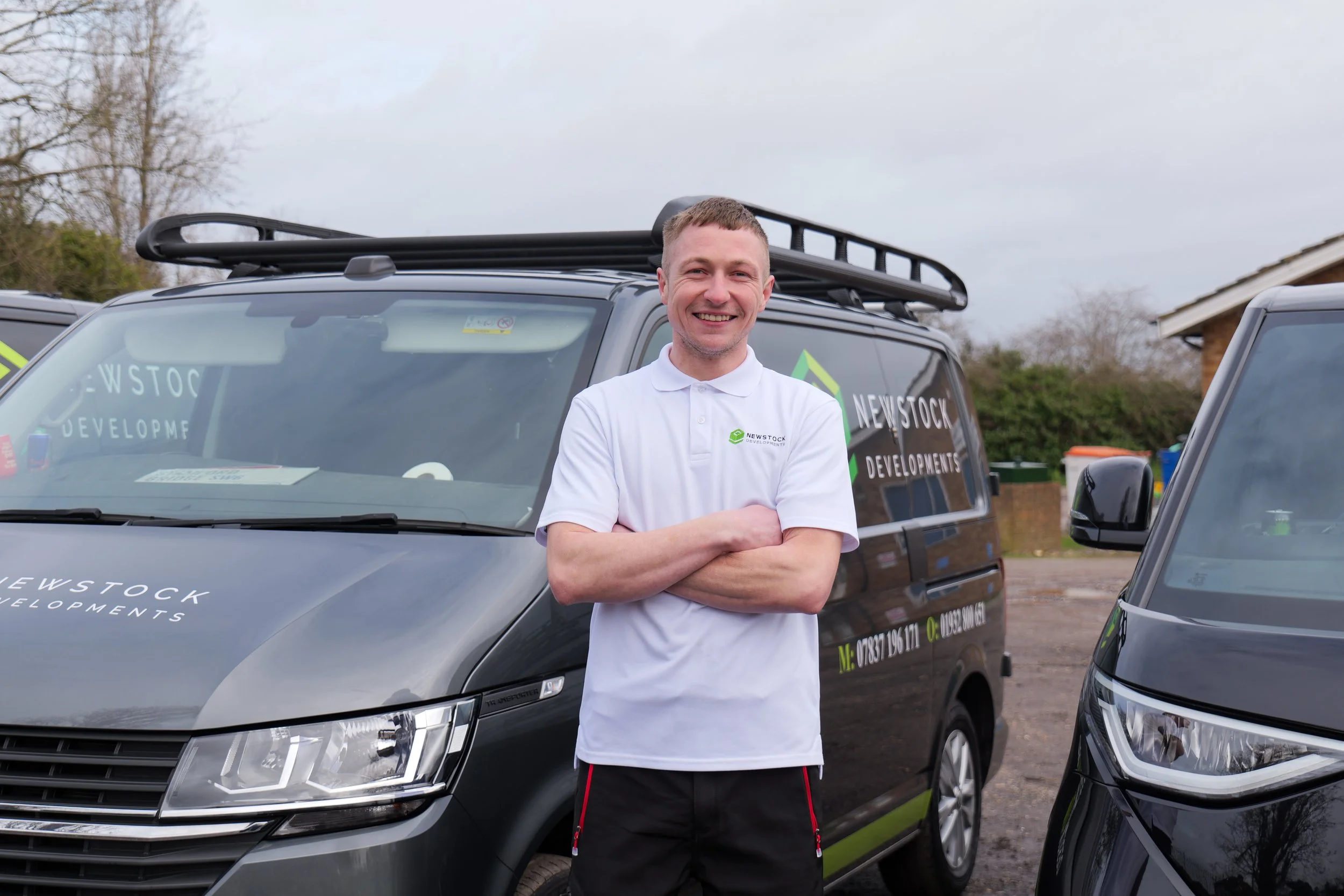 A smiling man standing outdoors in front of a grey vehicle with a logo that says 'Newstock Developments'. He has short light brown hair and is wearing a white polo shirt and black shorts with red accents. The background shows overcast skies, trees, and part of a house.