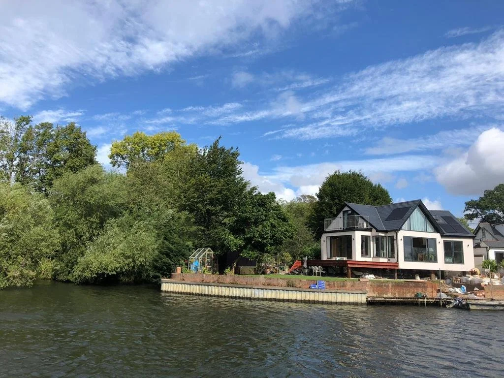 Modern house with large windows and a deck is situated along a river, surrounded by trees and a partly cloudy sky.