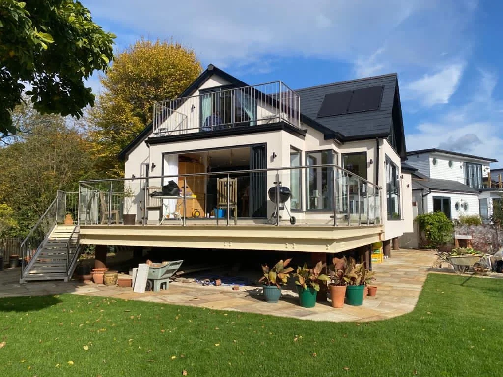 A modern house with a large wooden deck, black railing, and potted plants. The house has large glass windows and a black roof, surrounded by green lawn and trees under a partly cloudy sky.
