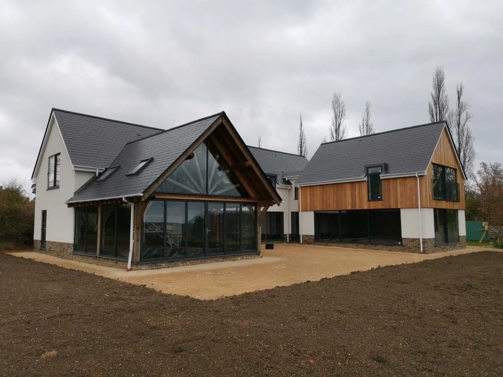 Modern house with a combination of white and wooden exterior, large glass windows, and a grey roof, situated on a plot with bare soil and no landscaping.