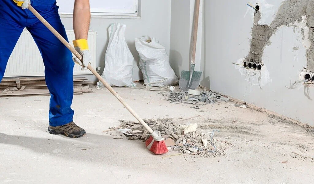 A person wearing blue work pants, gloves, and boots using a broom to sweep debris on a construction site inside a room with broken drywall and exposed wiring.