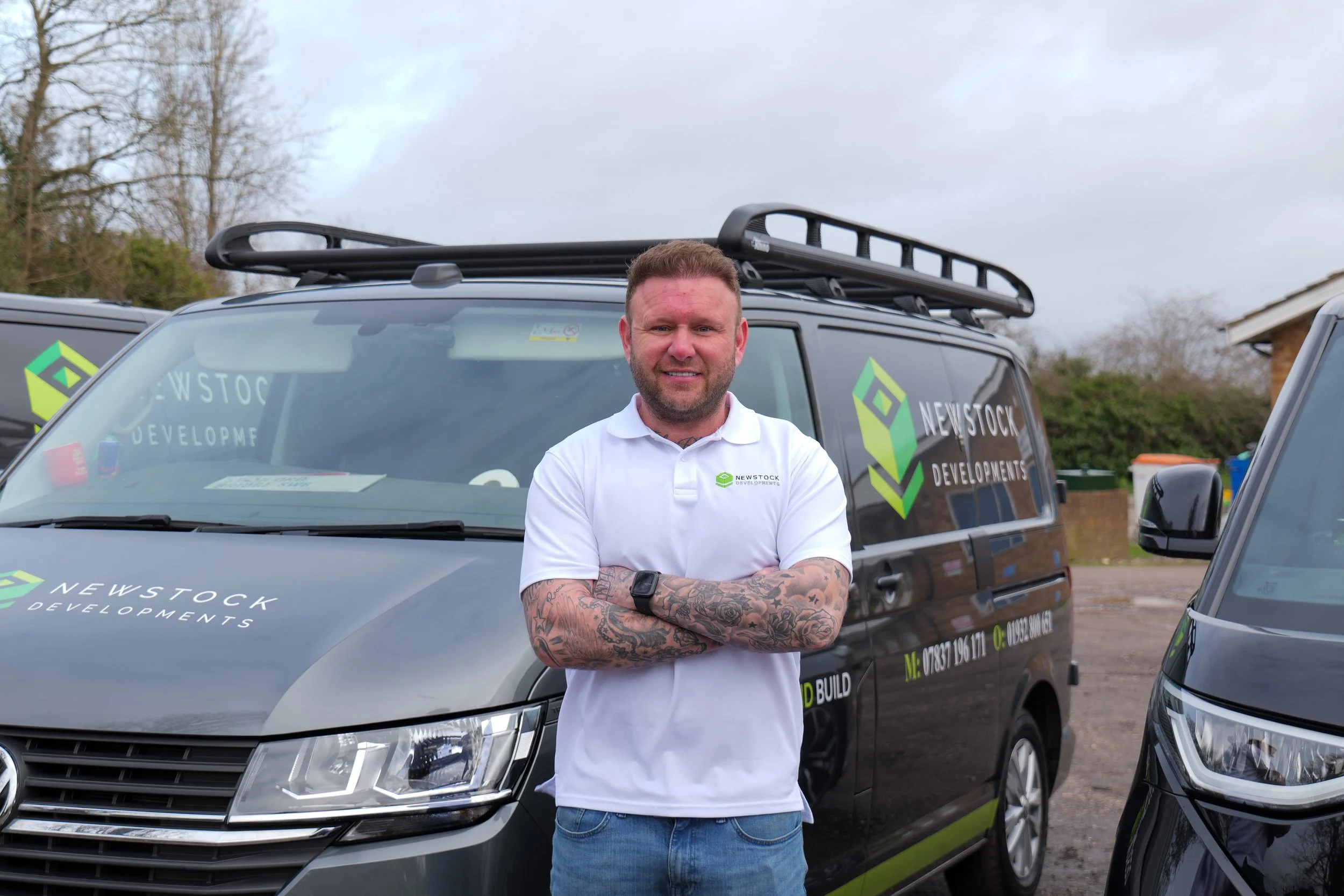 Man wearing a white polo shirt, standing with arms crossed in front of a black van with company logos and contact information, parked outdoors with trees and a cloudy sky in the background.