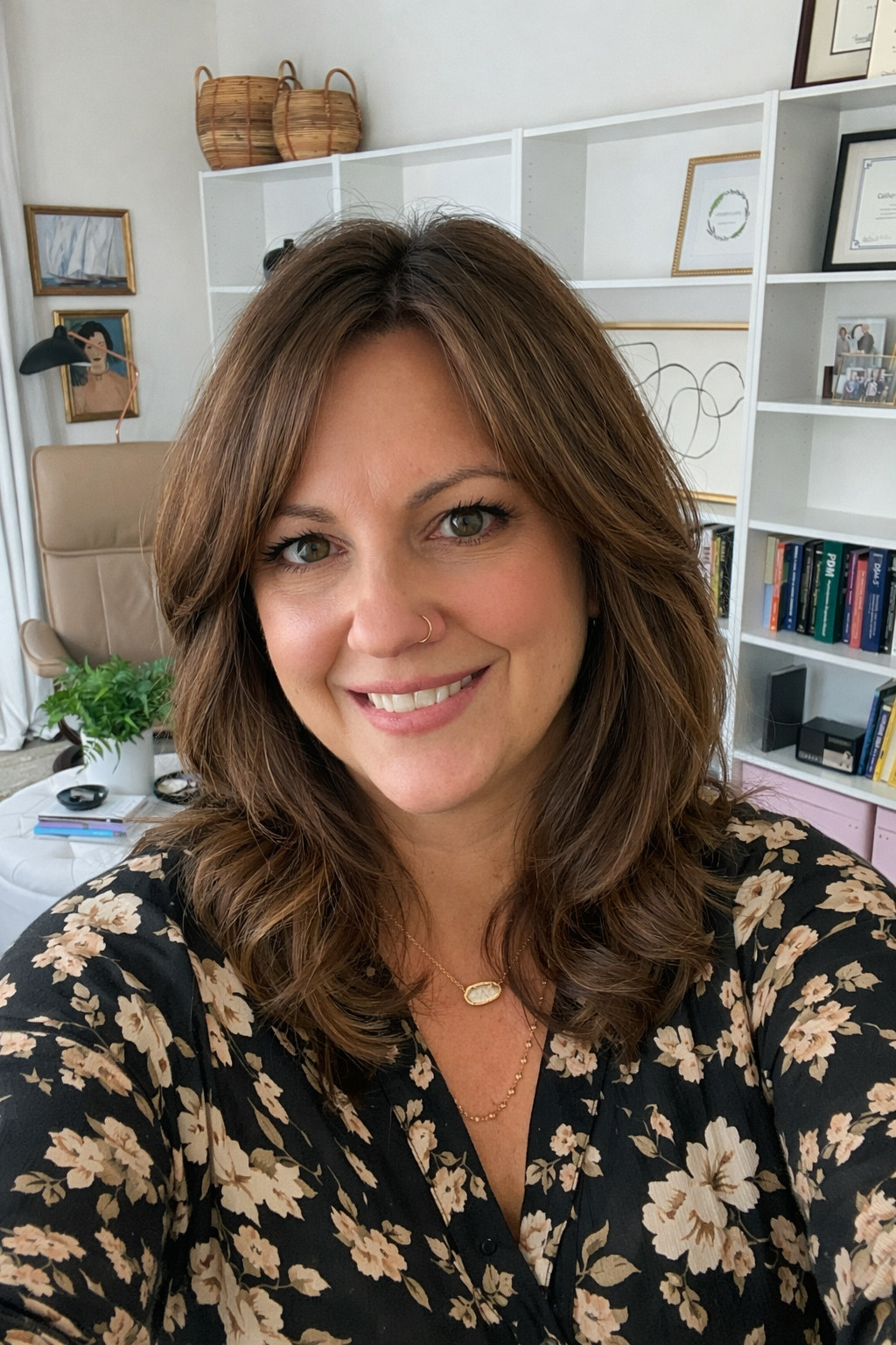 A woman with brown hair and a floral blouse smiling in an office with a white bookshelf, framed artwork, a plant, and certificates on the wall.