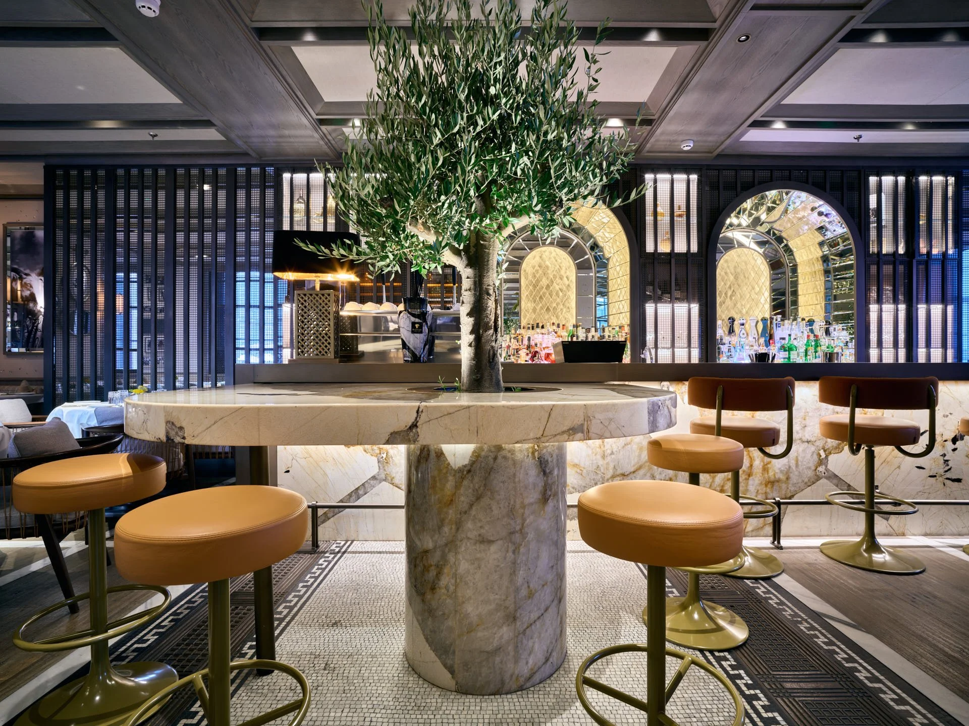 Circular stone table with an olive tree and cylindrical stone base in front of the illuminated Paper Moon bar.