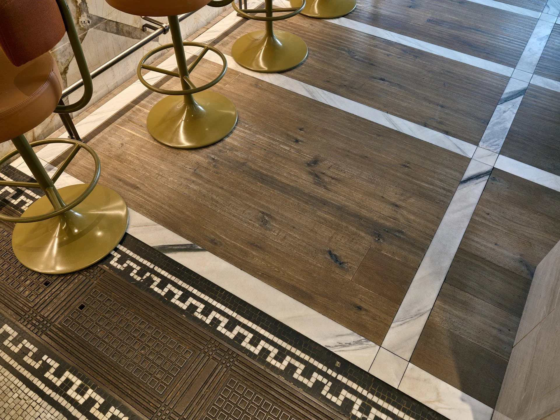 White marble bands inset into a timber floor beside black-and-white mosaic and cast-iron floor grilles.