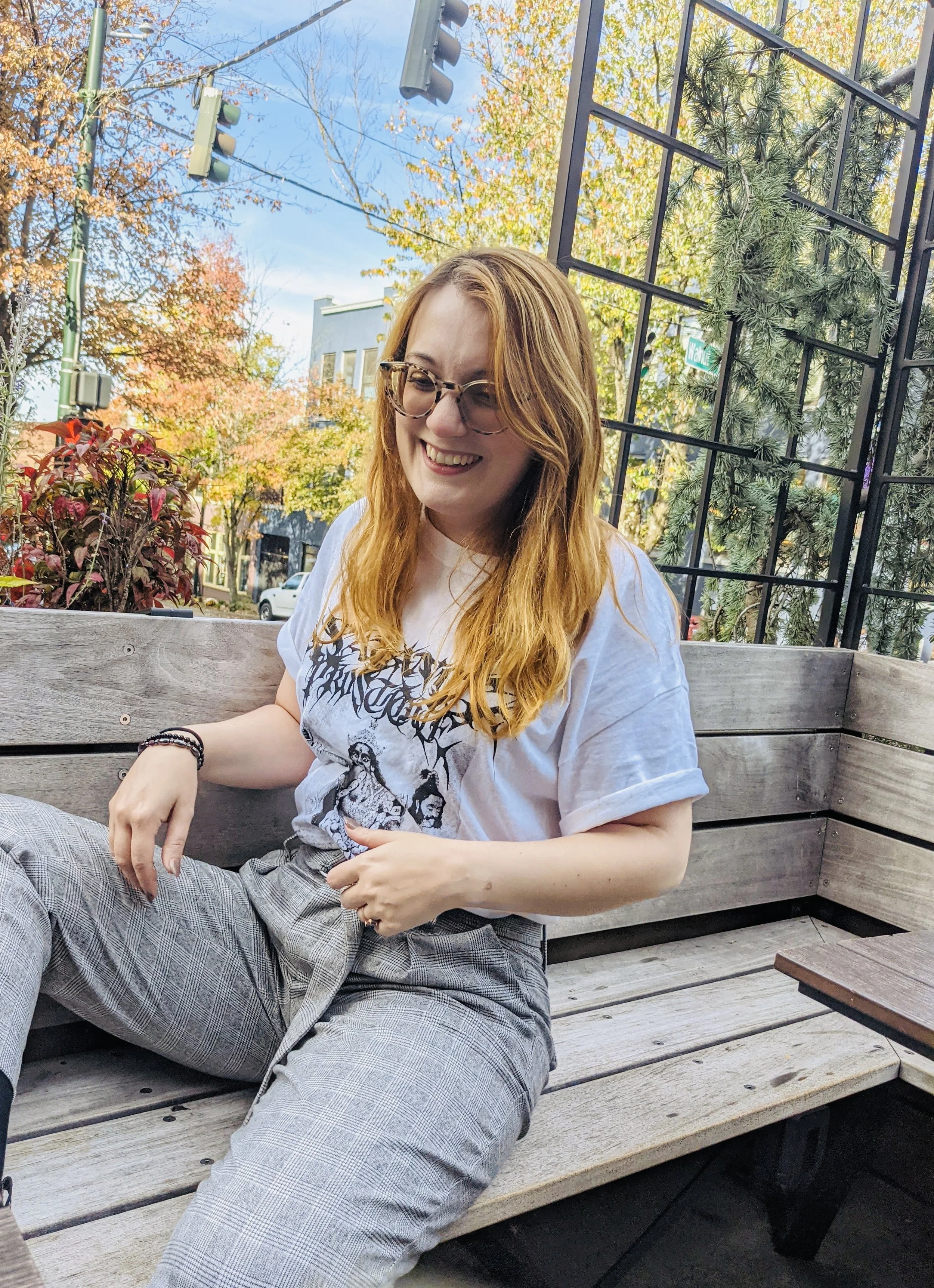A woman with long red hair, glasses, smiling, sitting on a wooden bench outdoors with autumn trees and a metal grid structure in the background.