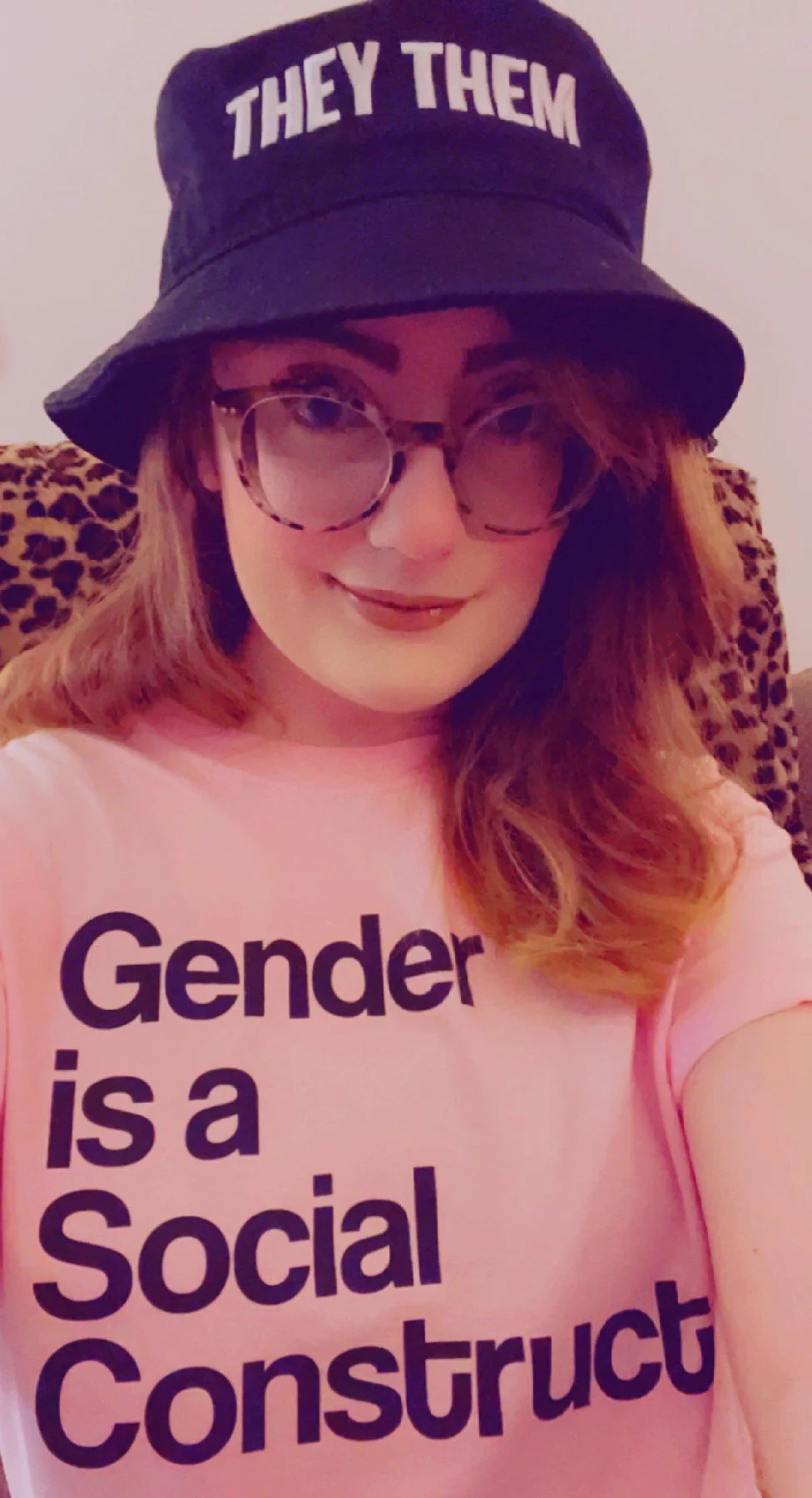 A woman wearing a pink shirt with bold black text that reads "Gender is a Social Construct', a black bucket hat with white embroidered text that reads 'THEY/THEM', and large tortoise-shell glasses. She is smiling and sitting on a leopard print chair.