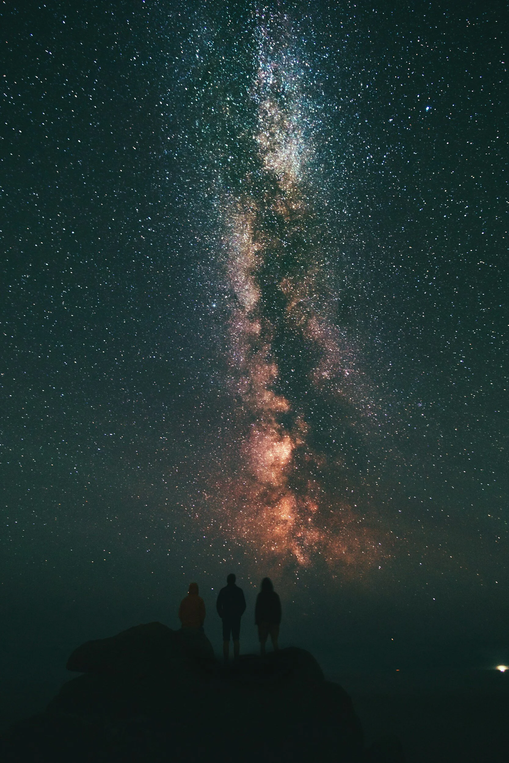 Three people standing on a rock formation, viewing the Milky Way galaxy in a clear, starry night sky.