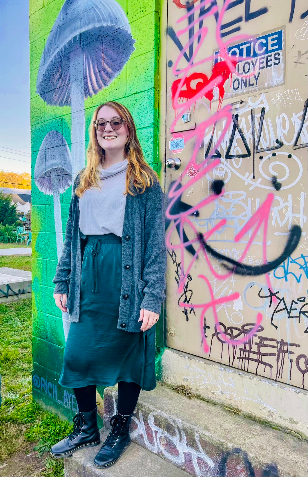 A young woman with glasses and long red hair stands smiling in front of a colorful graffiti mural on a brick wall. The mural features purple mushrooms against a bright green background. To her right, there is a door covered with various tags, graffiti, and stickers, including a sticker that reads 'NOTICE: Employees Only.' She is wearing a gray cardigan, light gray turtleneck, dark green skirt, black tights, and black combat boots, standing on a concrete sidewalk with green grassy area in the background.