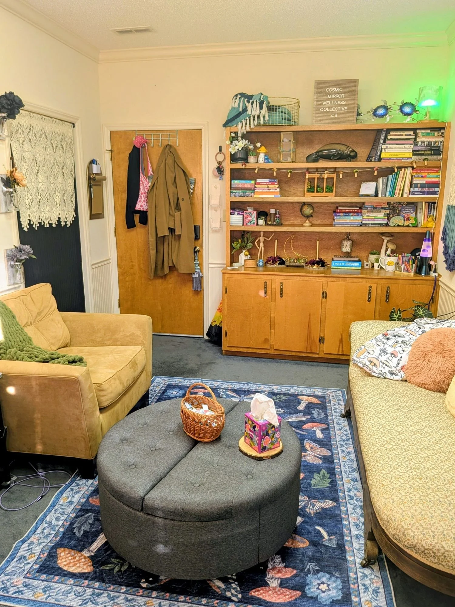 Living room with yellow and patterned sofas, a round ottoman with a basket and tissue box, a wooden bookshelf with books and decorative items, a front door with coats hanging, and a dark rug with floral and butterfly designs.