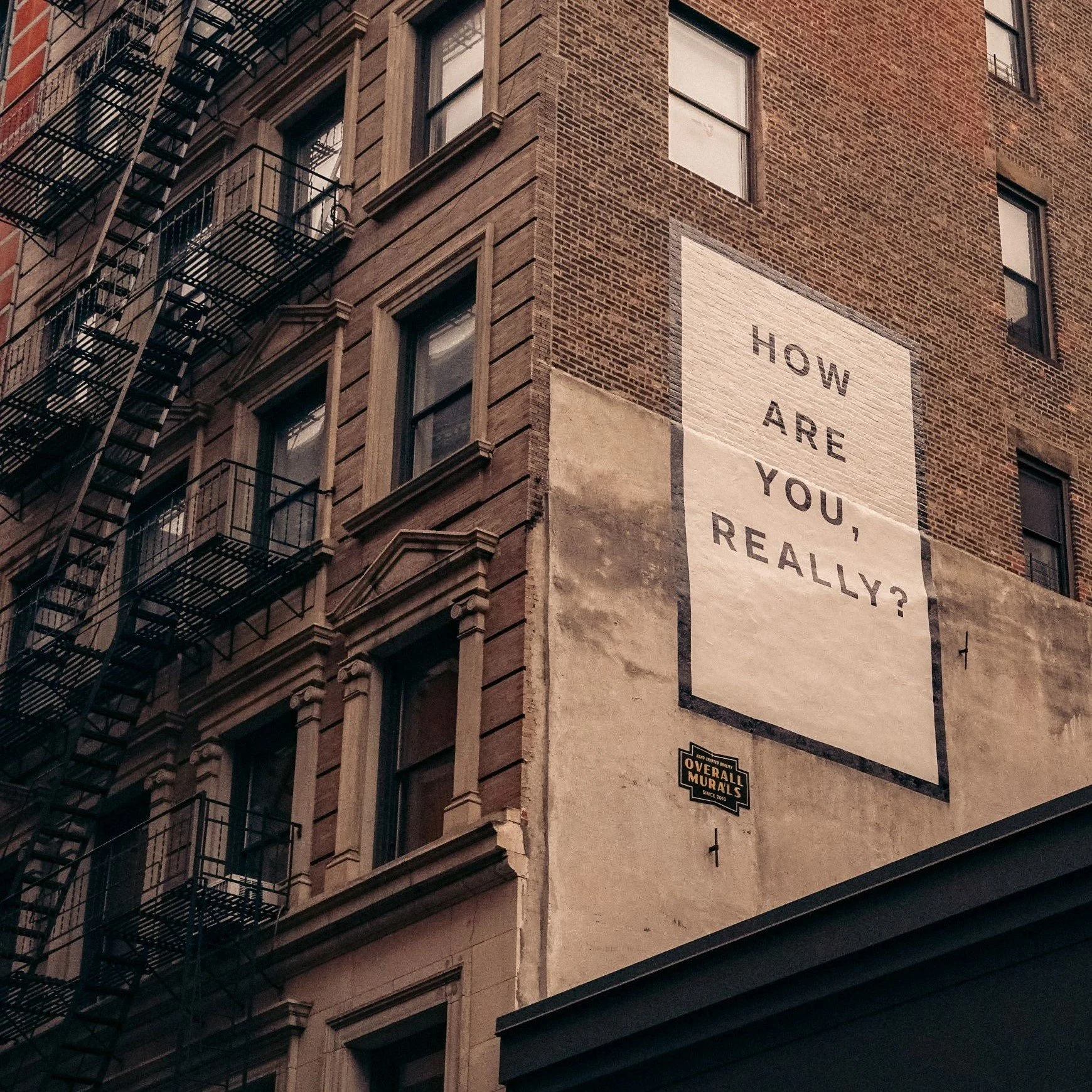 A large mural on the side of a brick building that reads, "How are you, really?" in black letters on a white background.