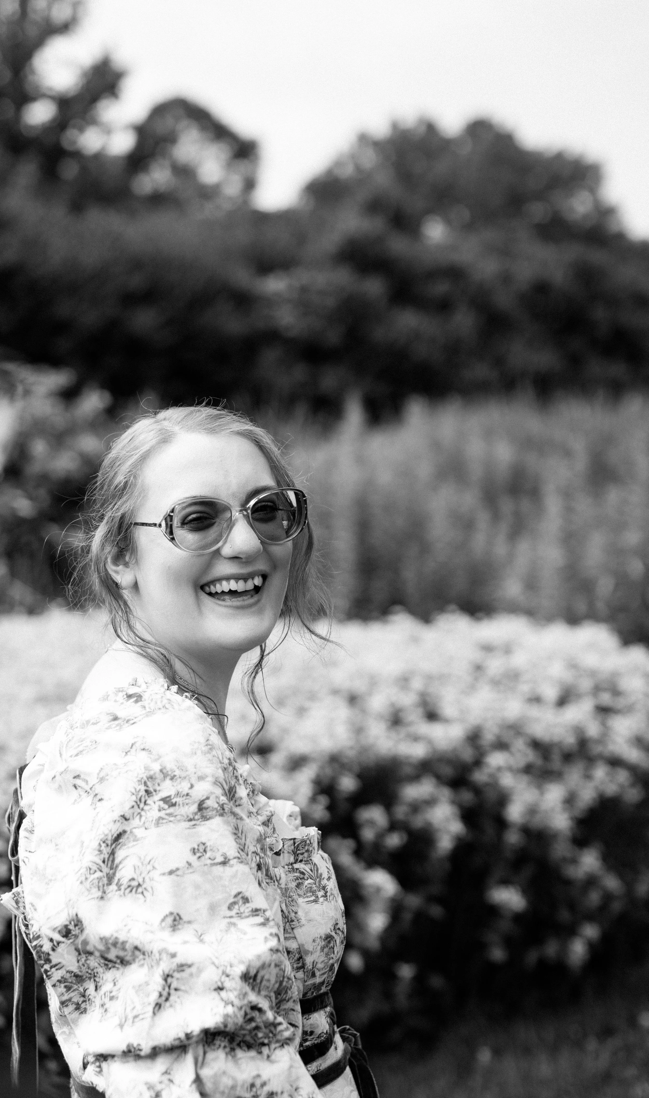 A woman wearing glasses and a floral blouse, smiling outdoors in what appears to be a garden or park, black and white photo.