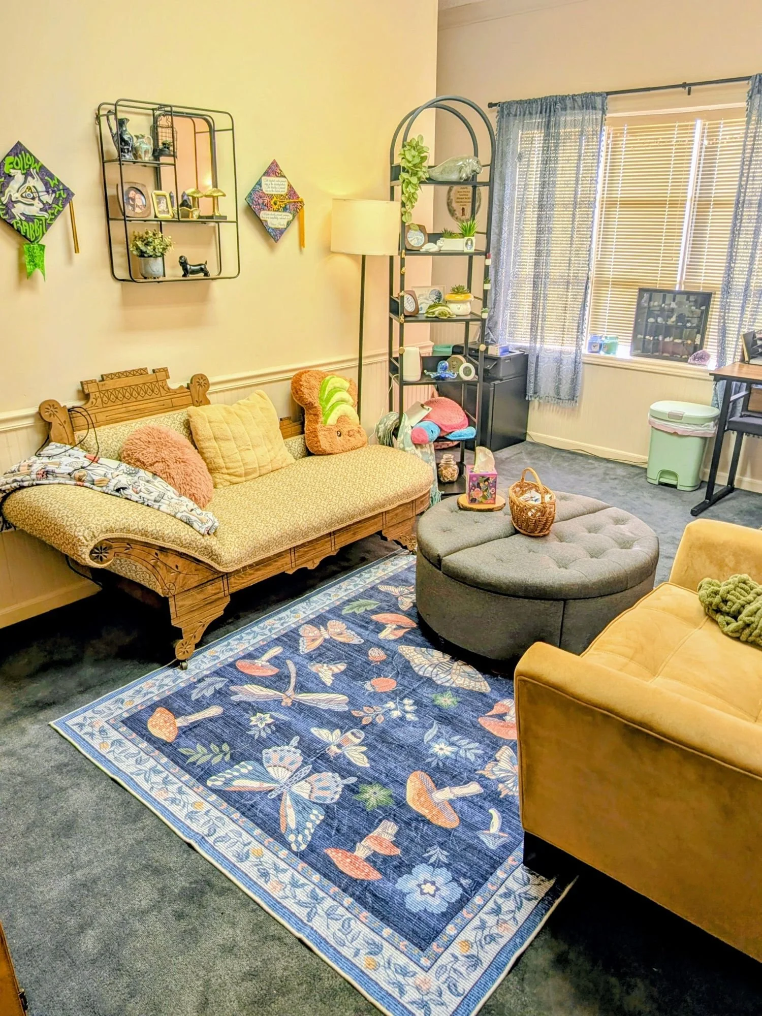 Living room with a patterned yellow sofa, yellow armchair, round ottoman, blue floral area rug, wall shelves with decor, a window with blinds and curtains, and various toys and items on shelves and floor.