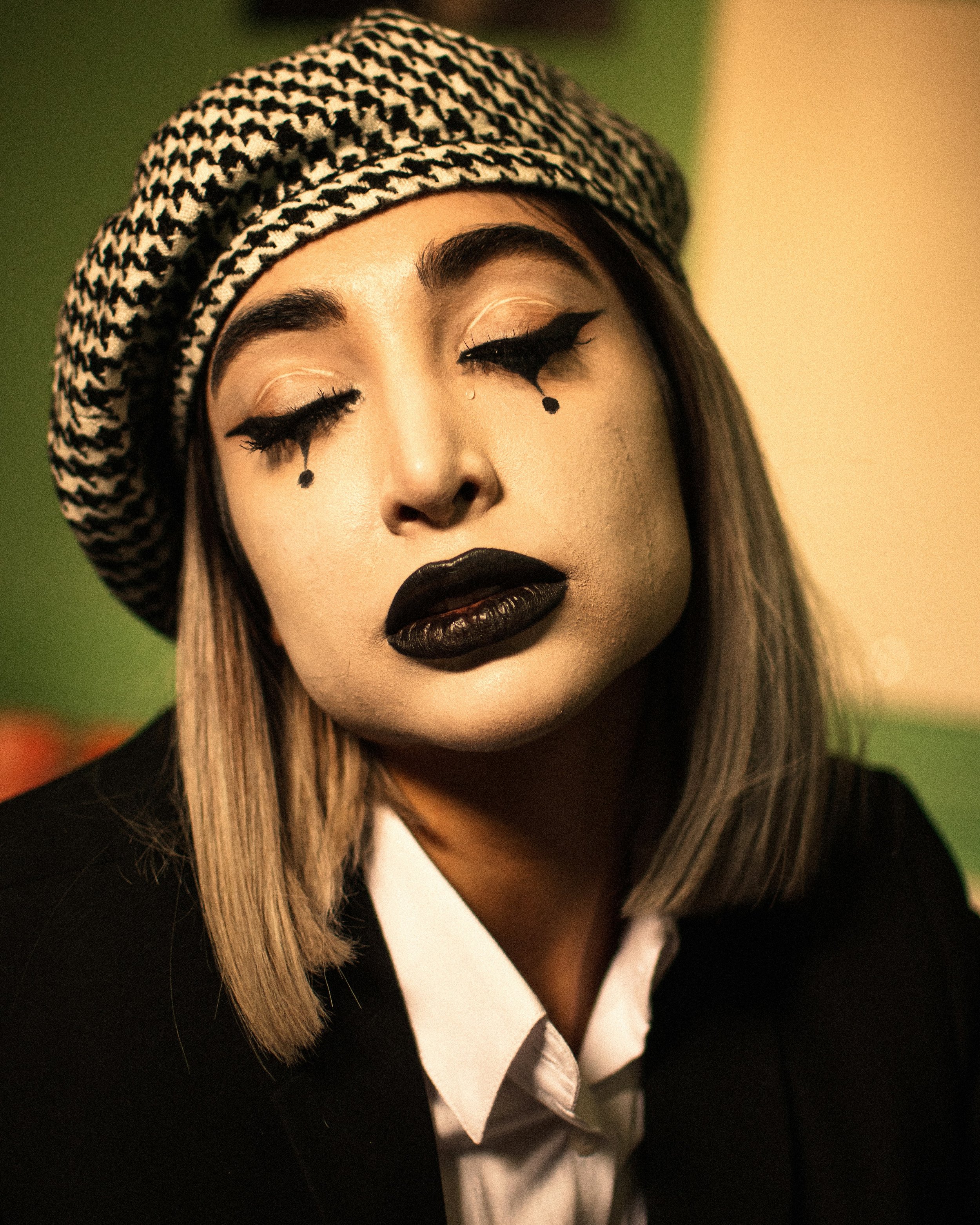 A person with blonde hair wearing a black and white patterned beret, with dark makeup including black lipstick and eyeliner, and artistic black tears drawn under closed eyes.