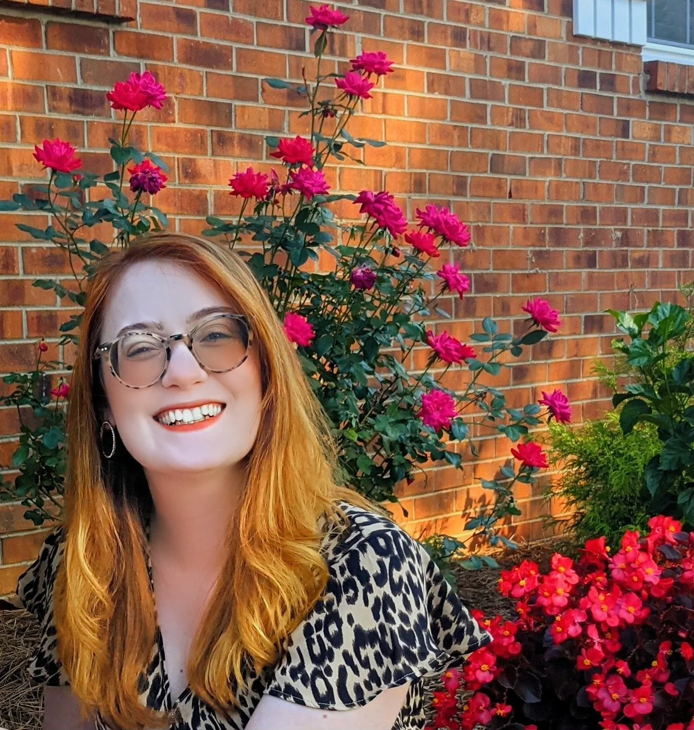 A woman with red hair, glasses, and a leopard print top smiling in front of blooming pink and red flowers near a brick wall.