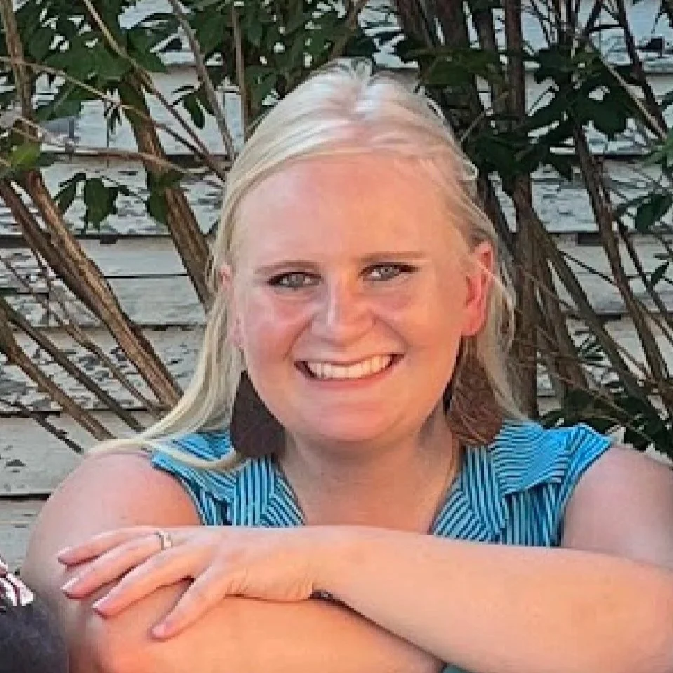 A woman with blonde hair and blue eyes smiling, wearing a blue striped top and large earrings, outdoors with greenery and a white wooden structure in the background.