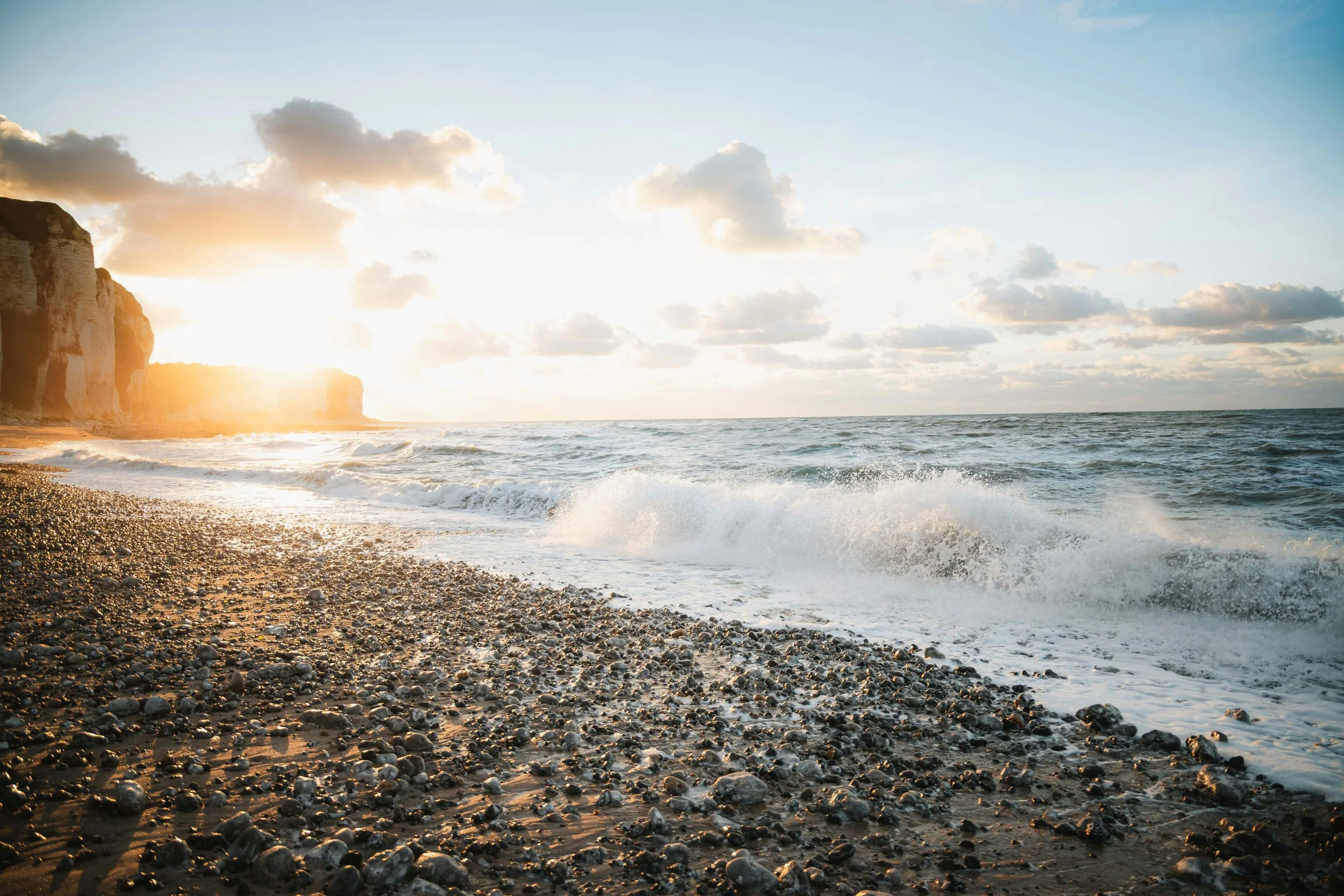 Sunset over a pebble beach with waves crashing and cliffs in the background