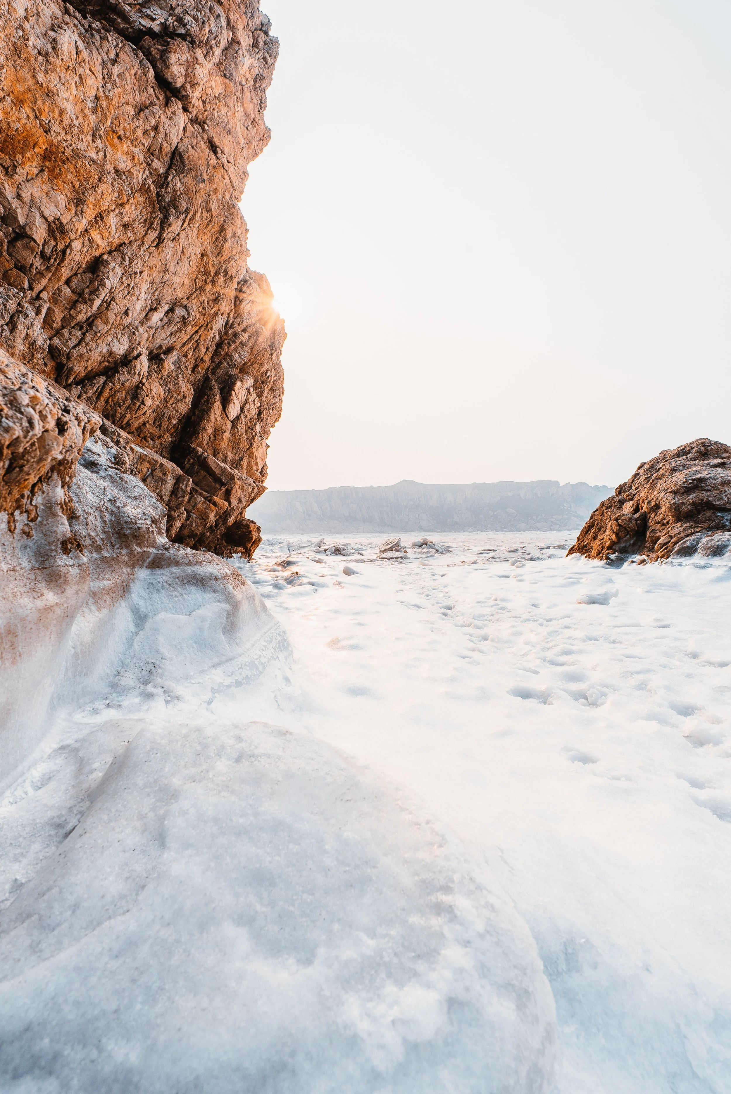A frozen coastal landscape with two large rugged rocks and ice covering the ground, with the sun peeking through the rocks and a distant shoreline in the background.