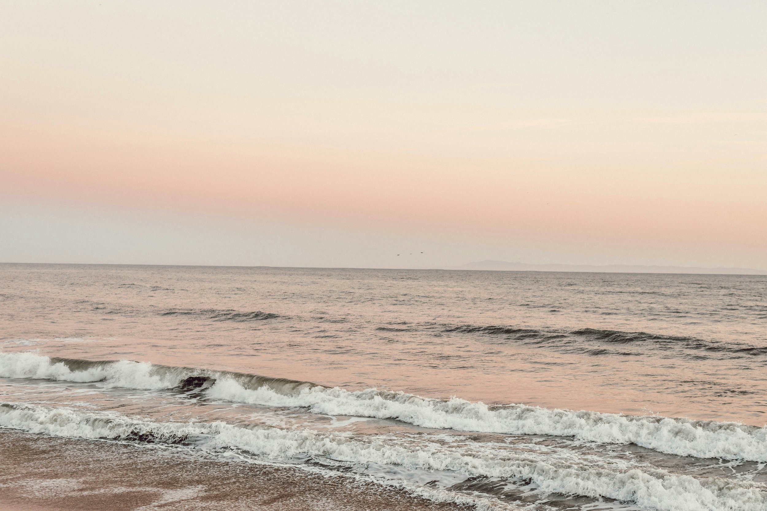 A serene beach scene with gentle waves approaching the shore during sunset, pastel-colored sky with pink, peach, and light blue hues, horizon in the distance, and faint landforms visible.